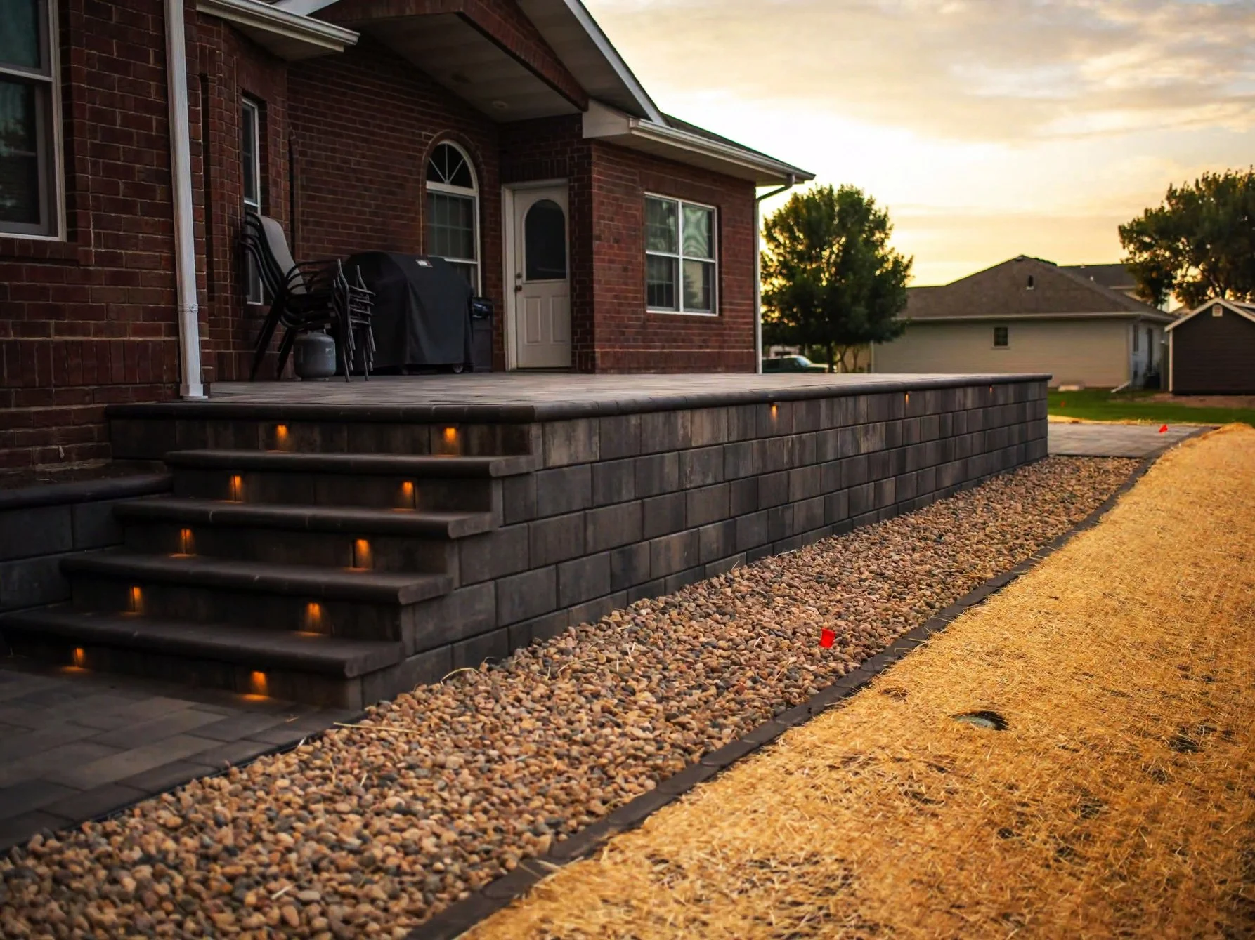Backyard patio area with a brick house, stairs with built-in lighting, and a gravel yard during sunset.
