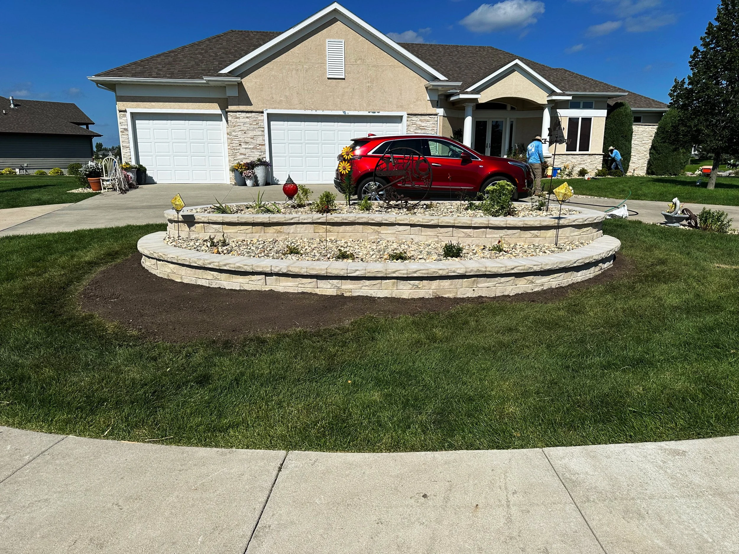 A residential house with a front yard that features a tiered stone wall garden with decorative plants and ornaments. A red car is parked in the driveway, and there are workers in blue uniforms on the lawn, possibly doing landscaping or yard work. The