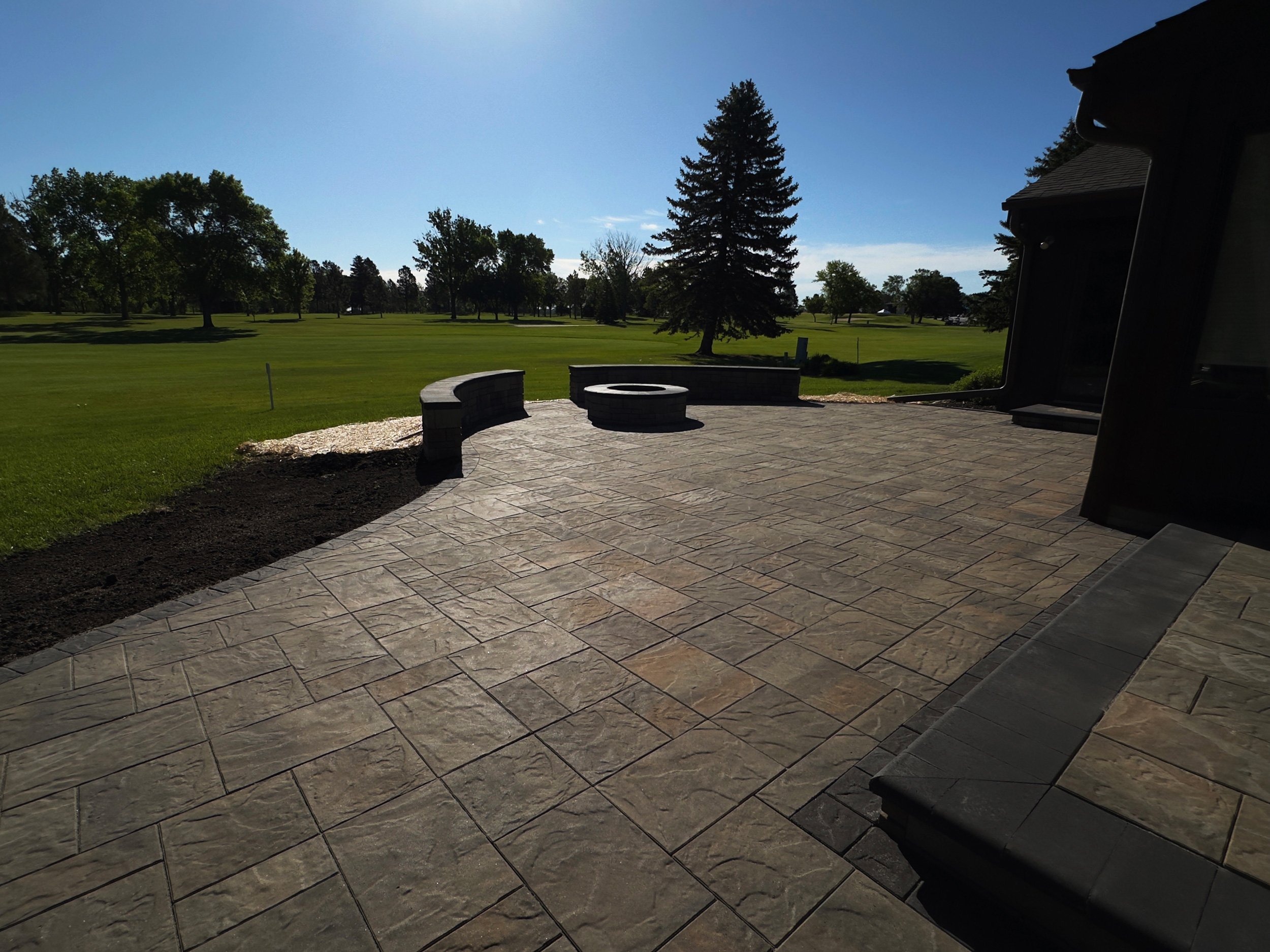 View of a backyard patio with stone flooring, curved stone benches, and a fire pit, overlooking a grassy field with trees under a clear blue sky.