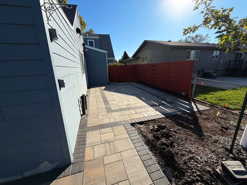 Backyard patio with paved stones and red wooden fence, adjacent to a light blue house, in a suburban neighborhood under clear blue sky.