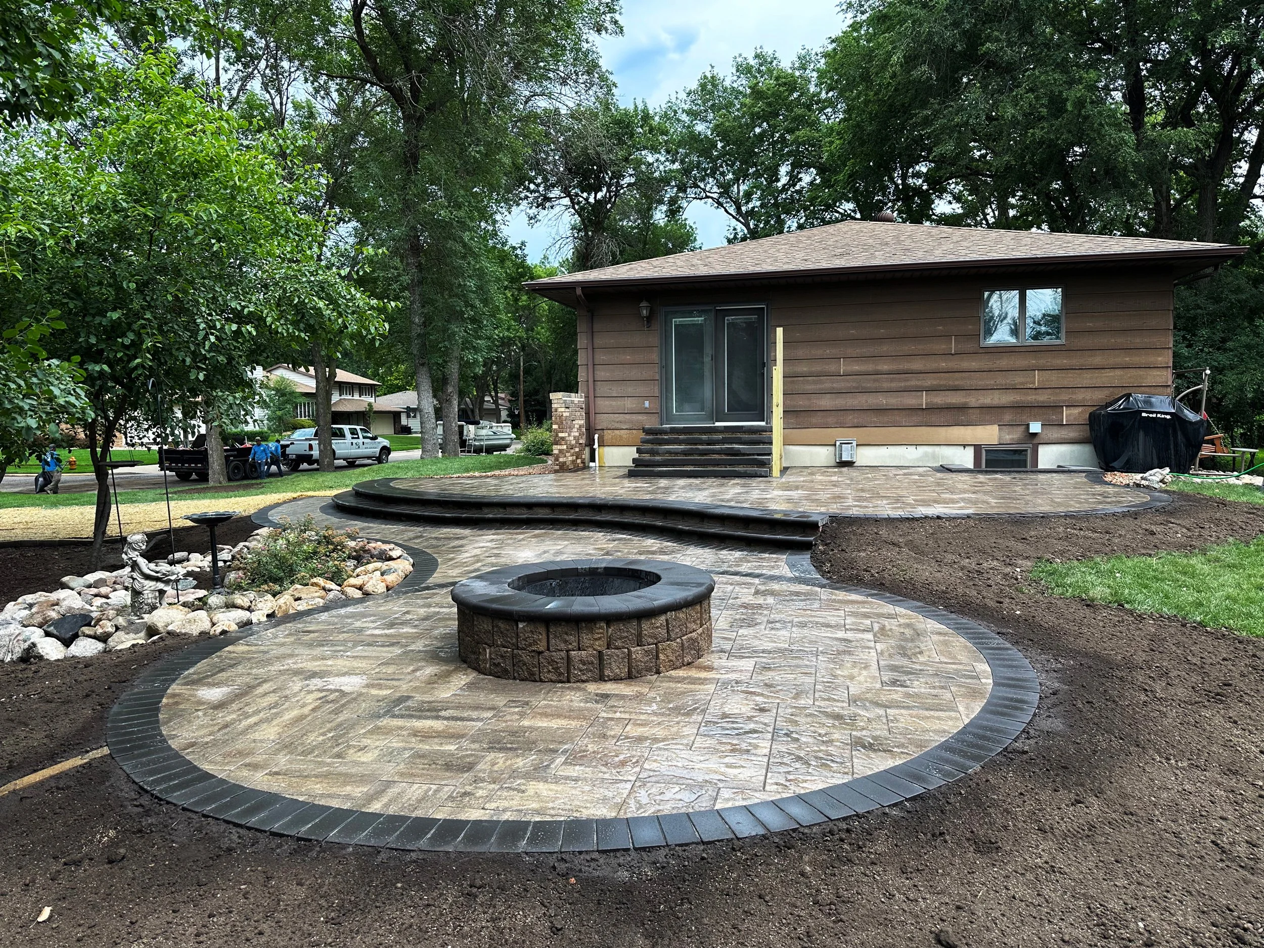 Backyard patio with a stone fire pit, curved steps, and a newly laid stone walkway leading to a house with brown wooden exterior and sliding glass door, surrounded by trees and green grass.