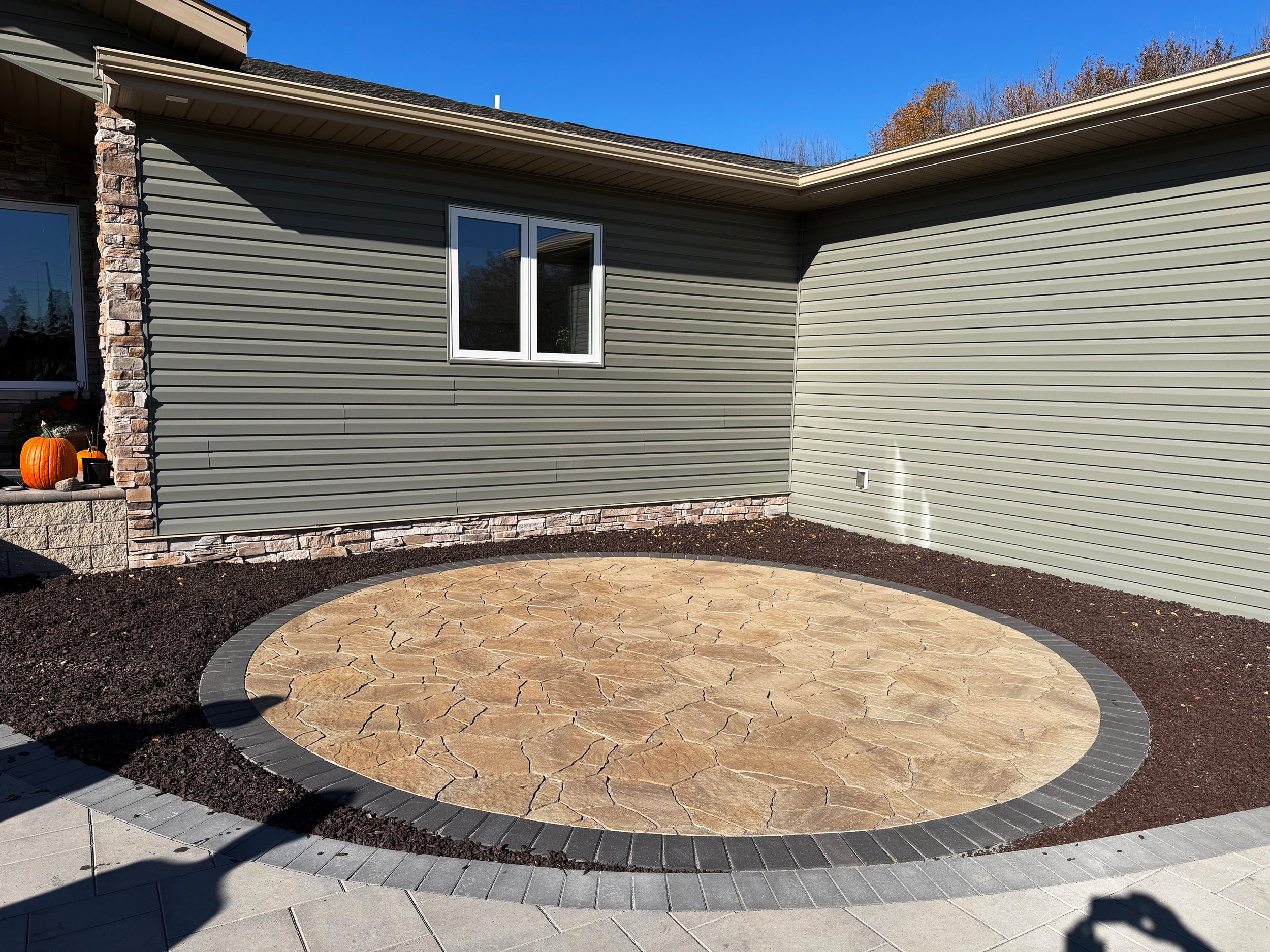 Circular paved patio area with stone pattern, bordered by gray bricks, adjacent to a house with gray siding and stone accents. Pumpkin decorations are visible on the porch.