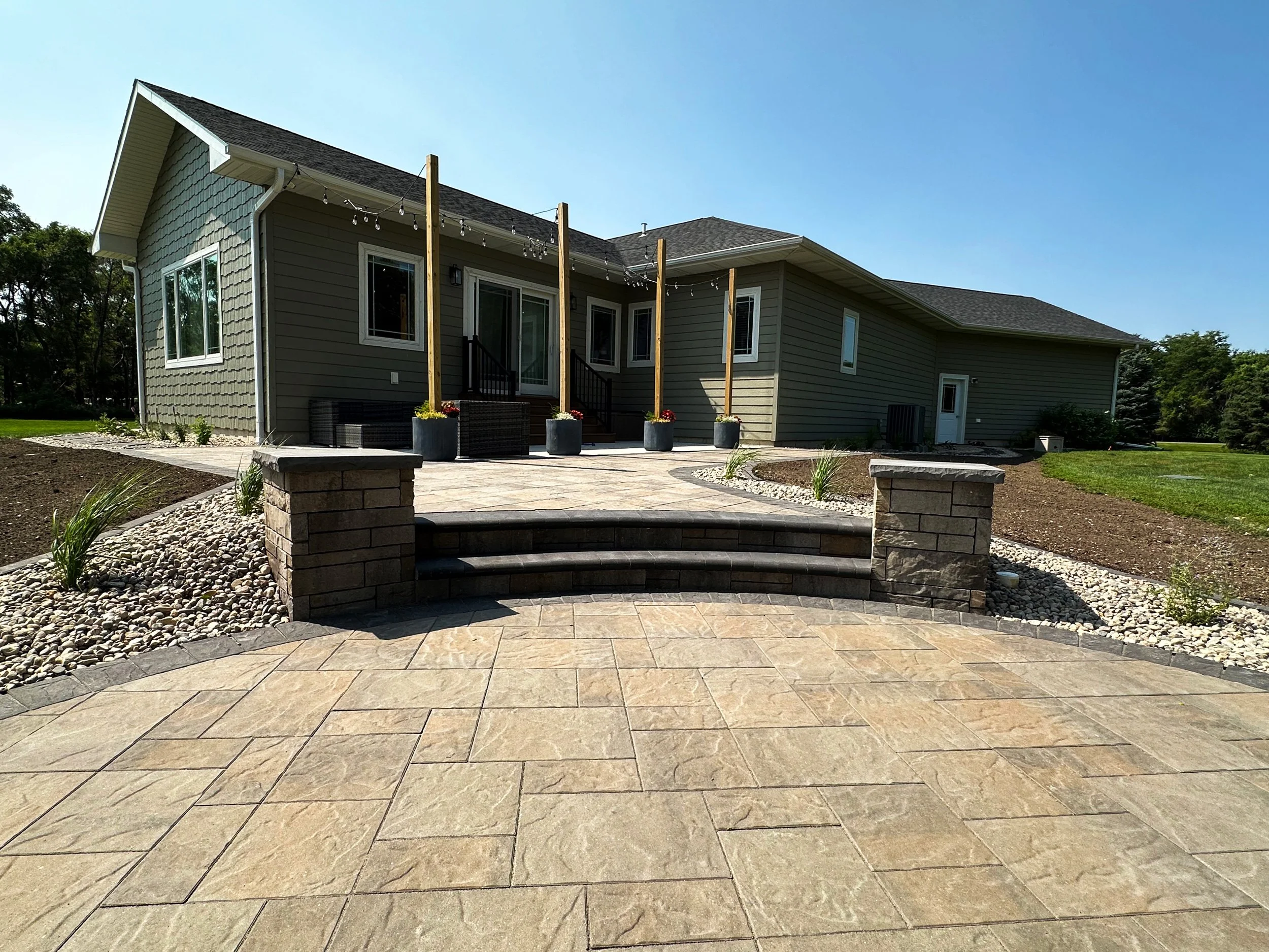 Backyard patio with stone tiles, steps, and landscaping, attached to a house with gray siding and a small deck with string lights.
