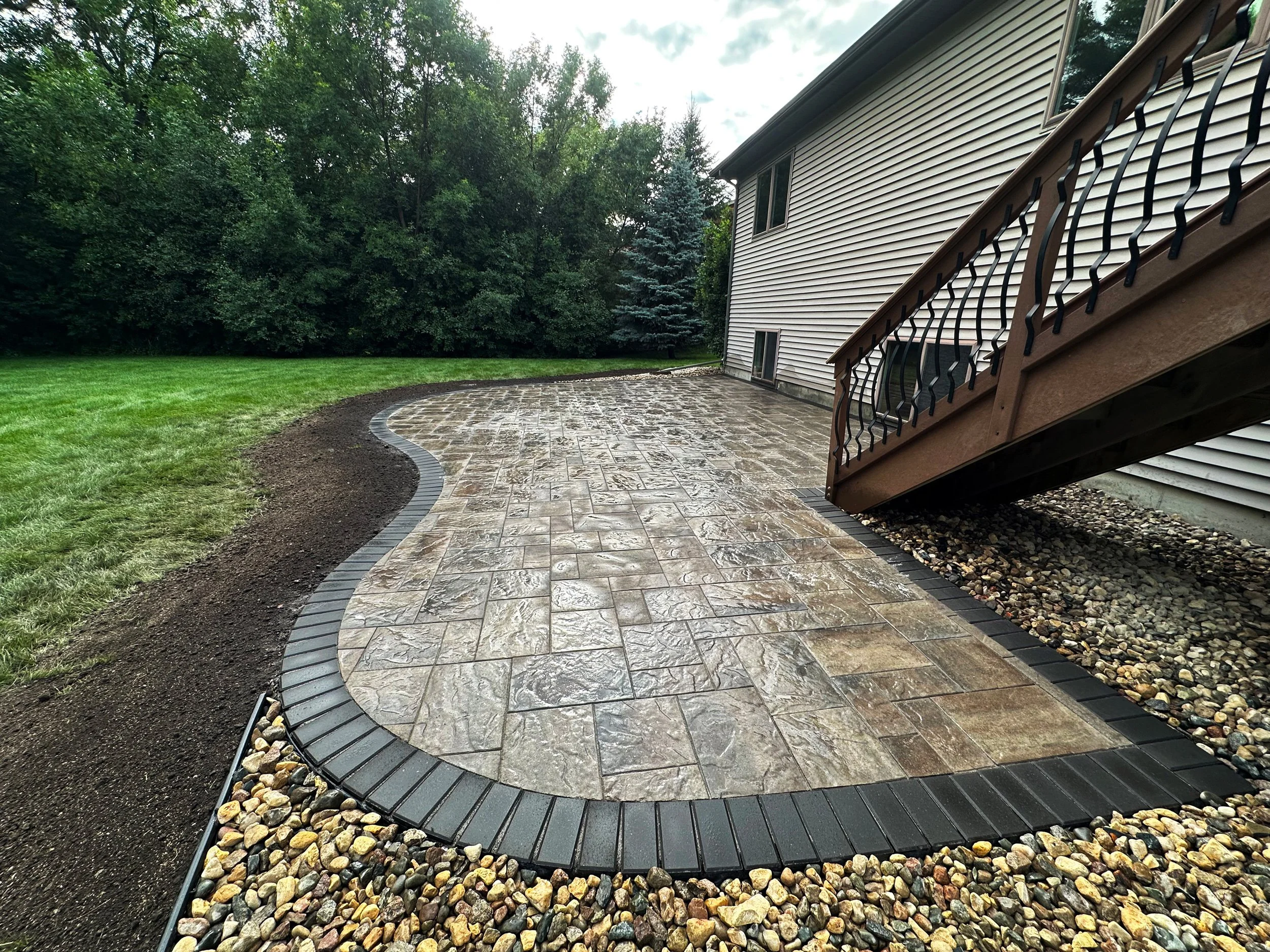 Freshly paved and bordered curved patio with decorative stone and brick edging next to a house with stairs, surrounded by a lawn and trees.