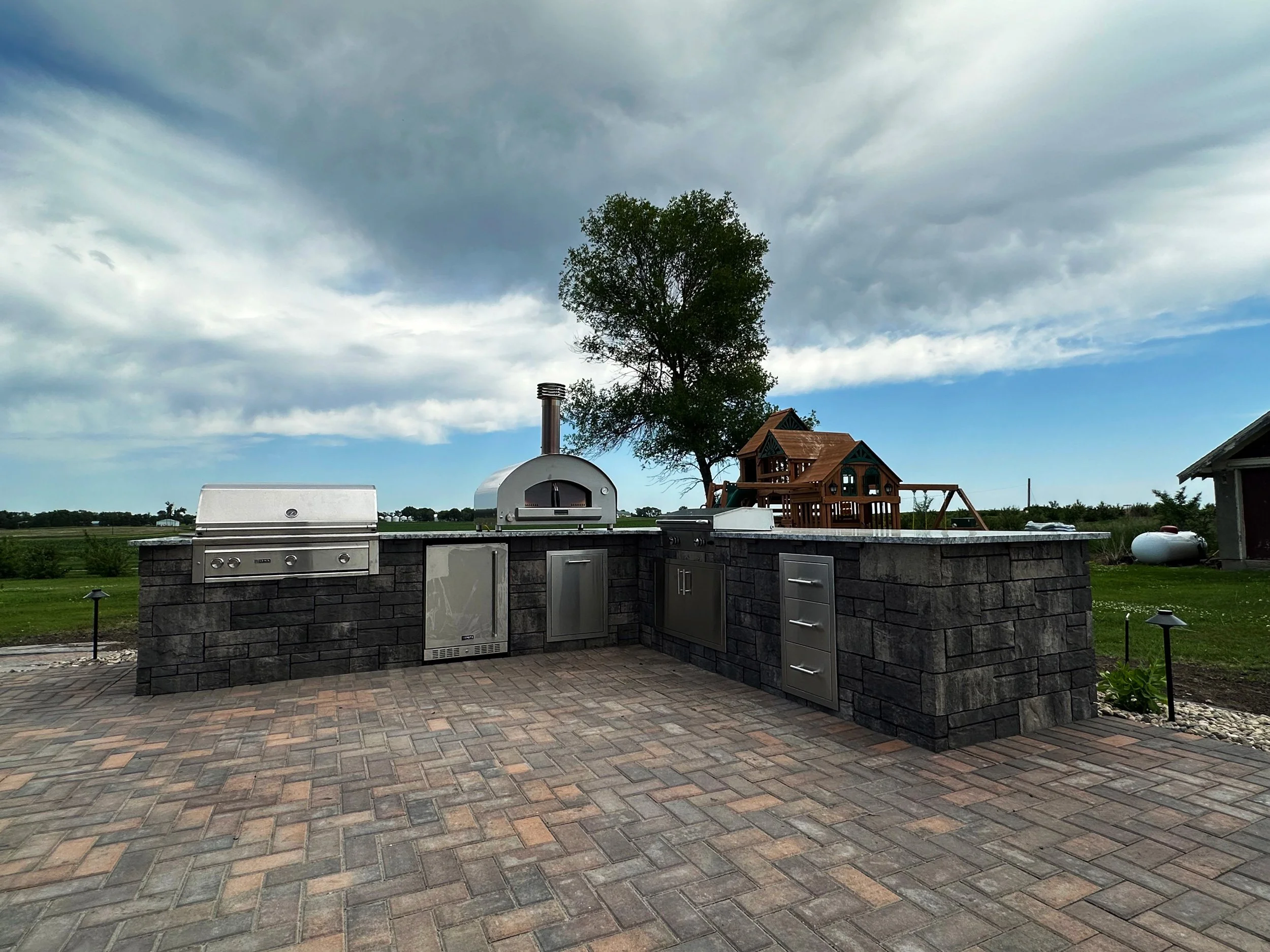 Outdoor kitchen with built-in grill, pizza oven, and storage cabinets on stone counter, with a brick patio, with a tree and wooden playset in the background under a partly cloudy sky.