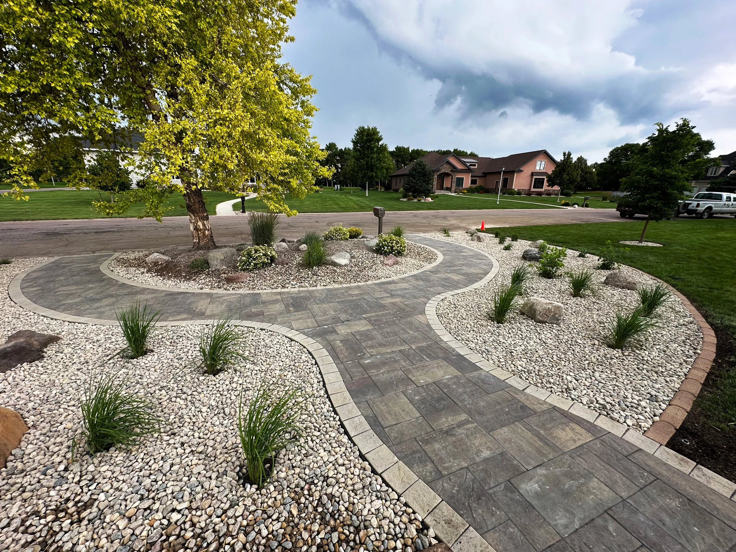 A landscaped yard featuring a curved stone pathway, a large tree, small plants, and rocks, with a residential neighborhood and cloudy sky in the background.
