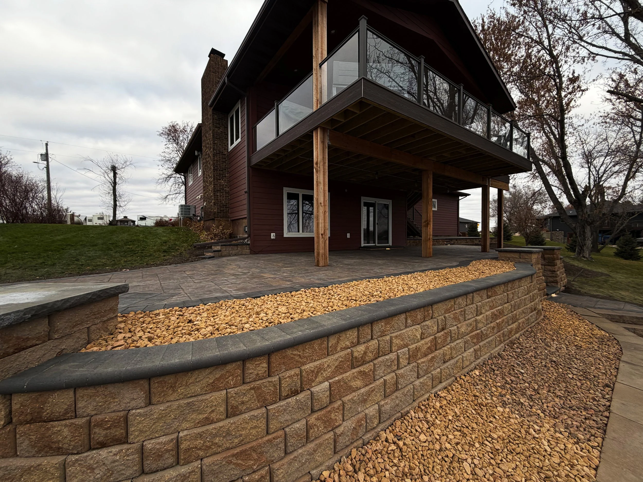 Backyard patio with stone retaining wall, gravel, and a two-story house with a balcony and sliding glass doors, under cloudy sky.