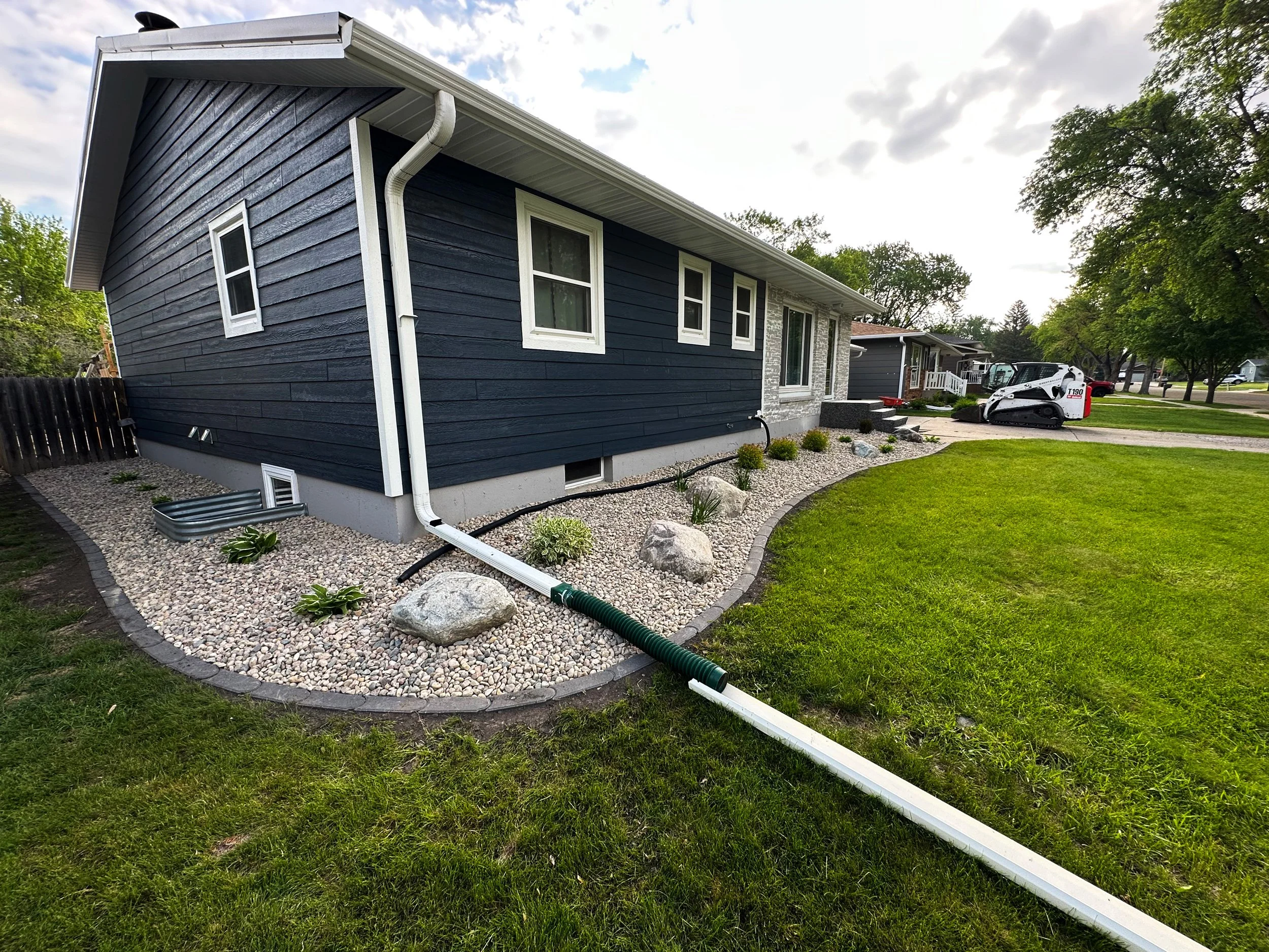 Side yard of a house with dark blue siding, white trim, and a rock garden with plants, a lawn, and a downspout extending from the roof to a drainage pipe. In the background, there is a sidewalk, trees, and a small street with parked cars, including a