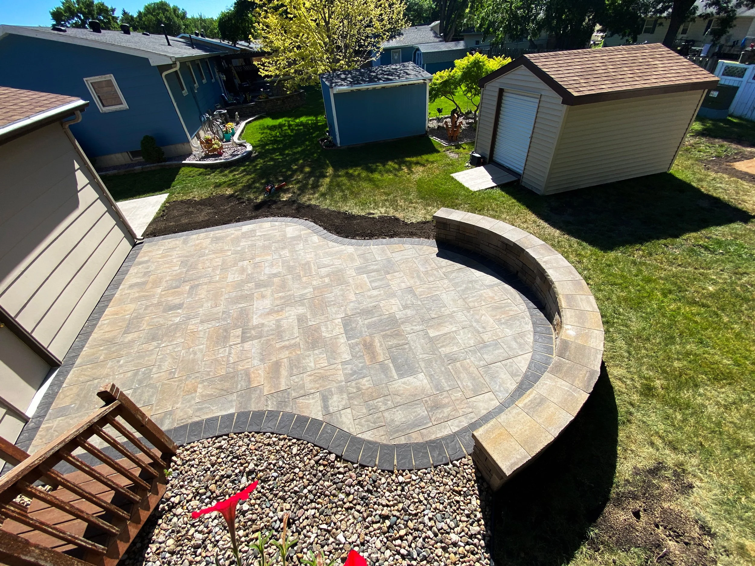 Backyard with a newly constructed stone patio, a small gravel area with red flowers, and additional structures including a beige shed, a blue shed, and a beige garage. There is green grass, trees, and neighboring houses visible in the background.