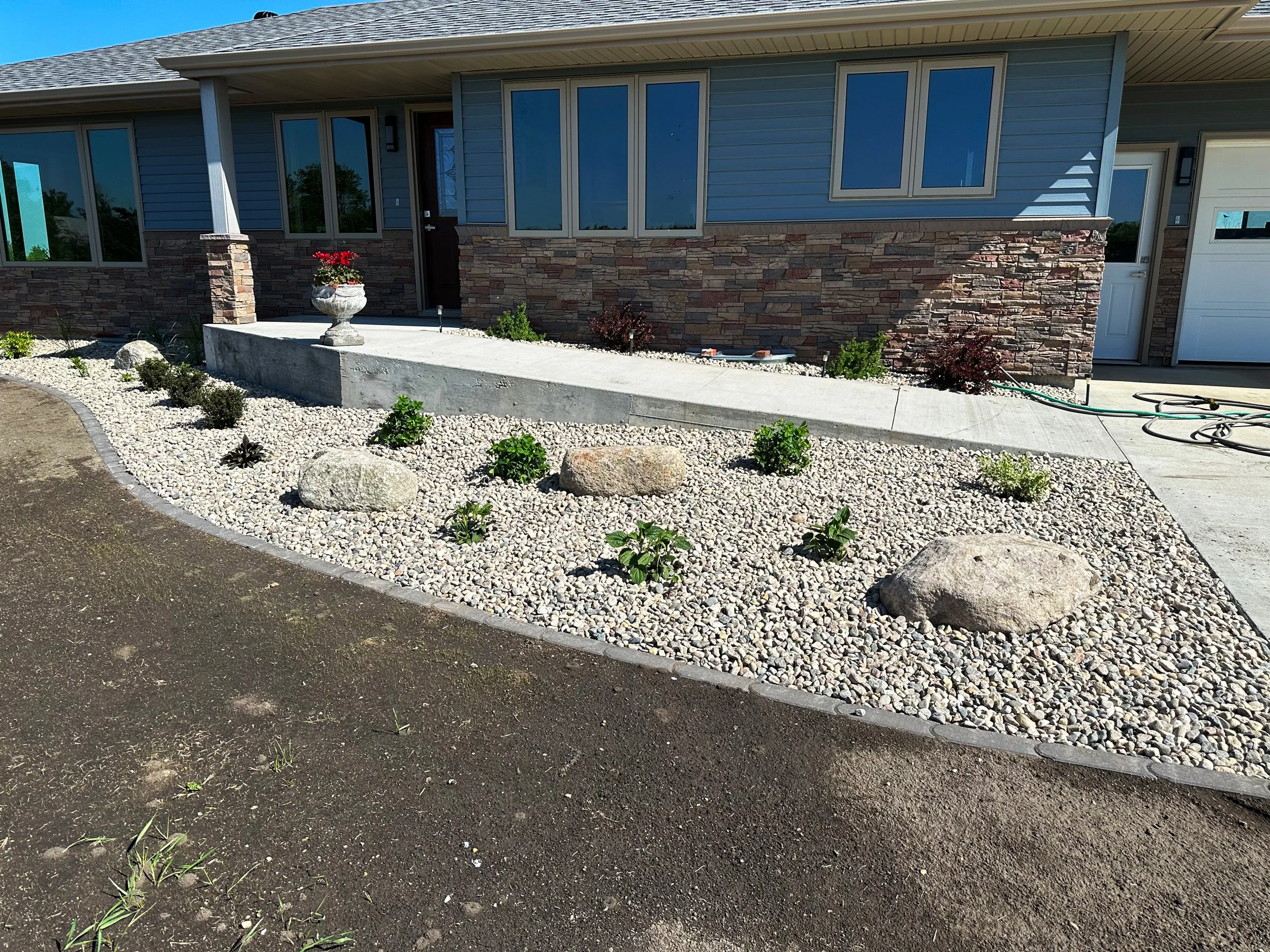 Front yard landscaping with a gravel bed, large stones, small green shrubs, a concrete pathway, and a house with blue siding, stone accents, and a white garage door.