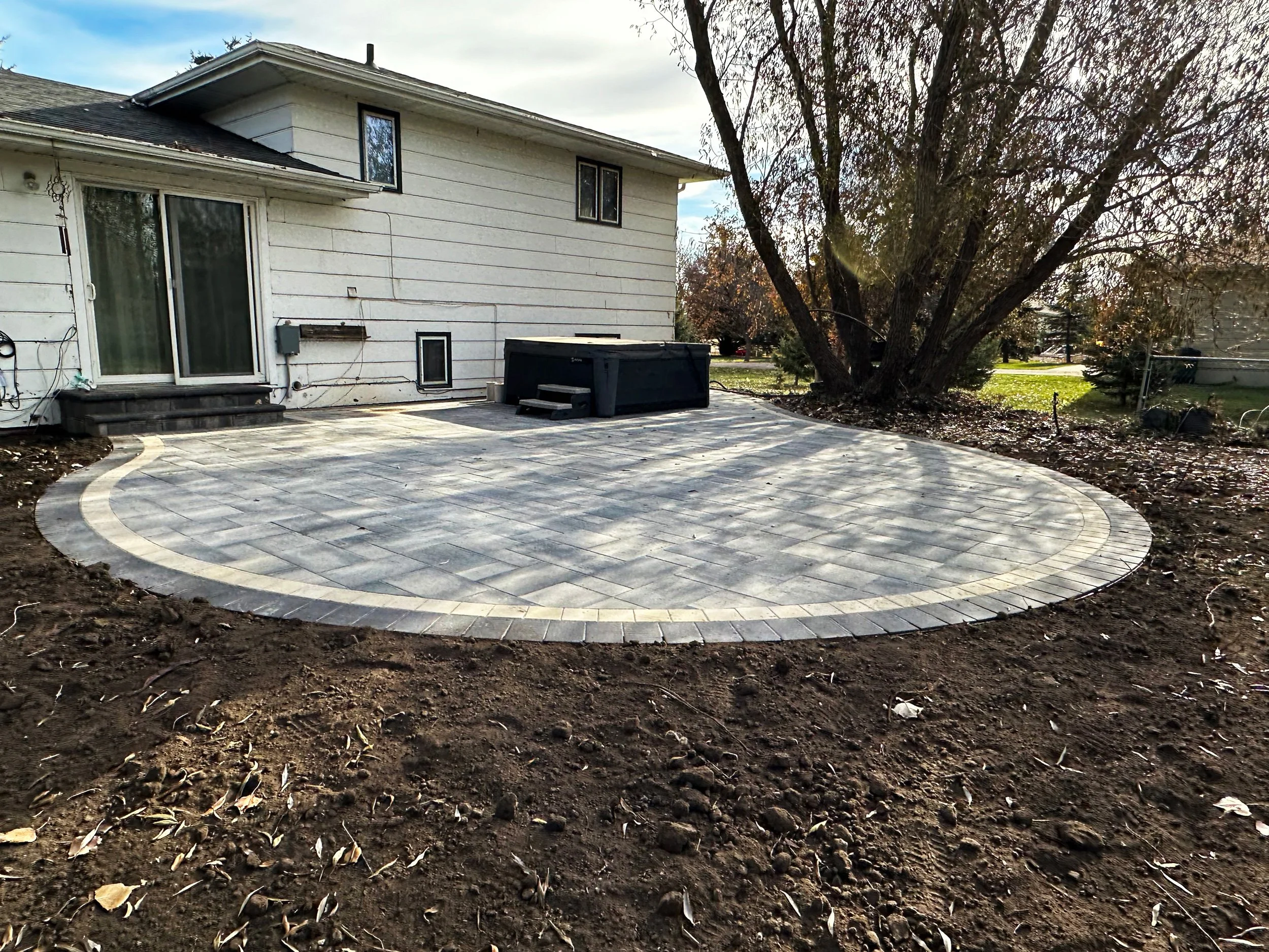 Backyard patio with a circular stone pattern, attached to a white house, with a large tree on the right and a grassy lawn in the background.