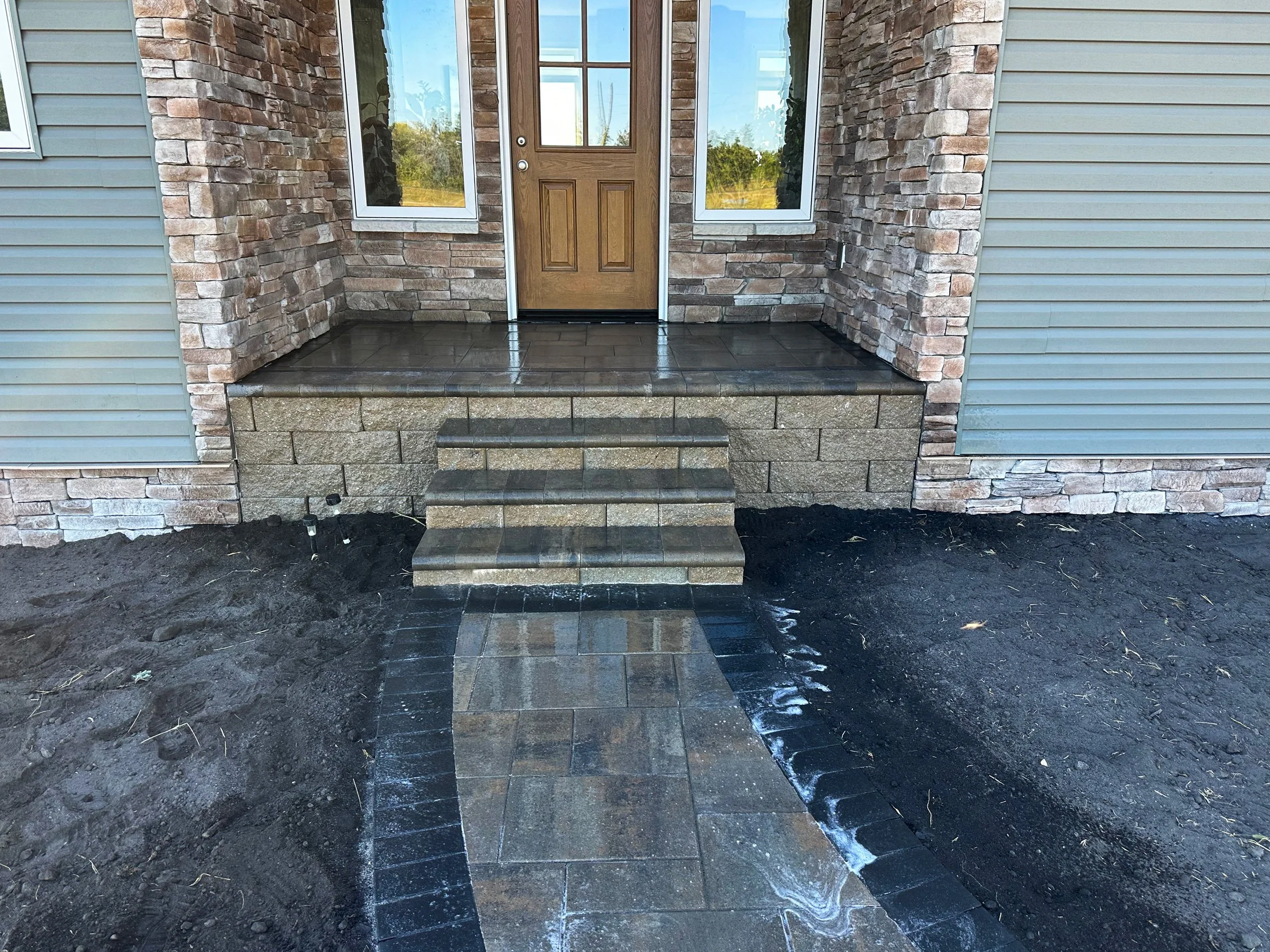 Front porch with stairs, brick and stone facade, wooden door, and steps leading up to door with surrounding paving and dirt ground.