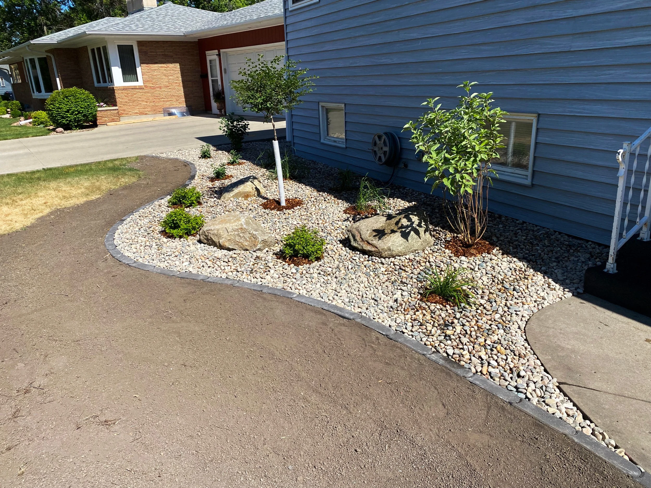 Front yard with landscaped garden bed featuring small shrubs, rocks, and mulch, bordered by stone edging, with a light blue house wall and a staircase on the right.