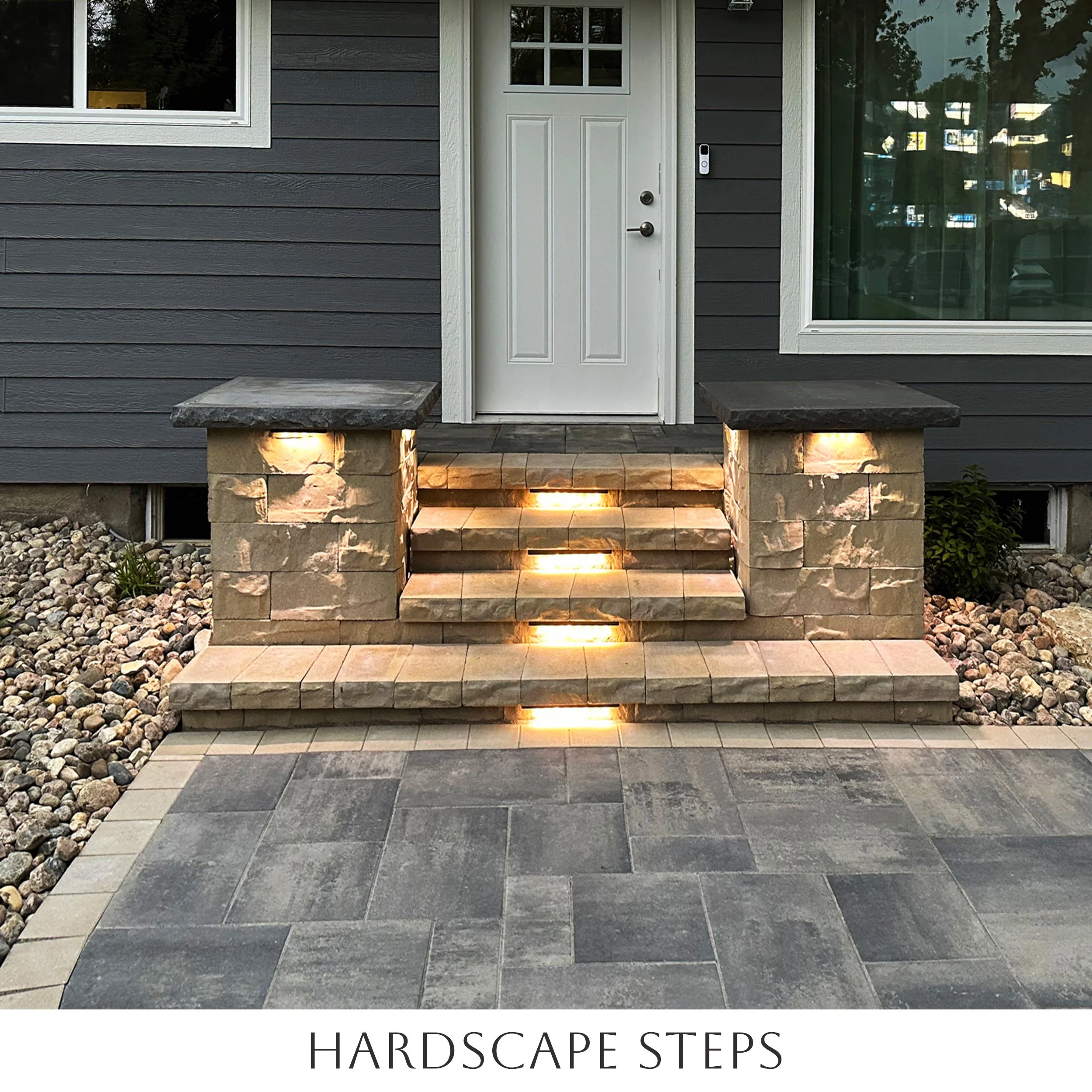 Exterior view of a house with a small staircase made of bricks, leading to a white front door. The stairs are lit with built-in lights, and the house has gray siding. There are landscaped rocks on either side of the stairs.