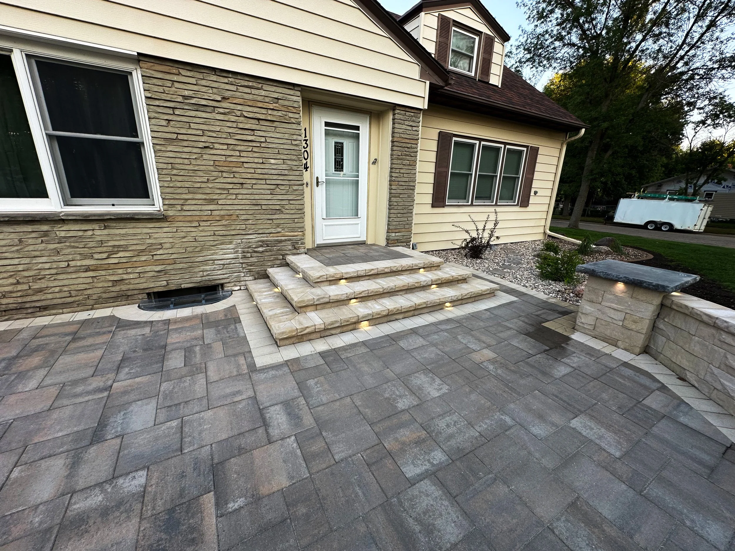 Front entrance of a house with stone steps leading to a door, surrounded by a paved patio area with outdoor lighting, and landscaped garden with small bushes and rocks.