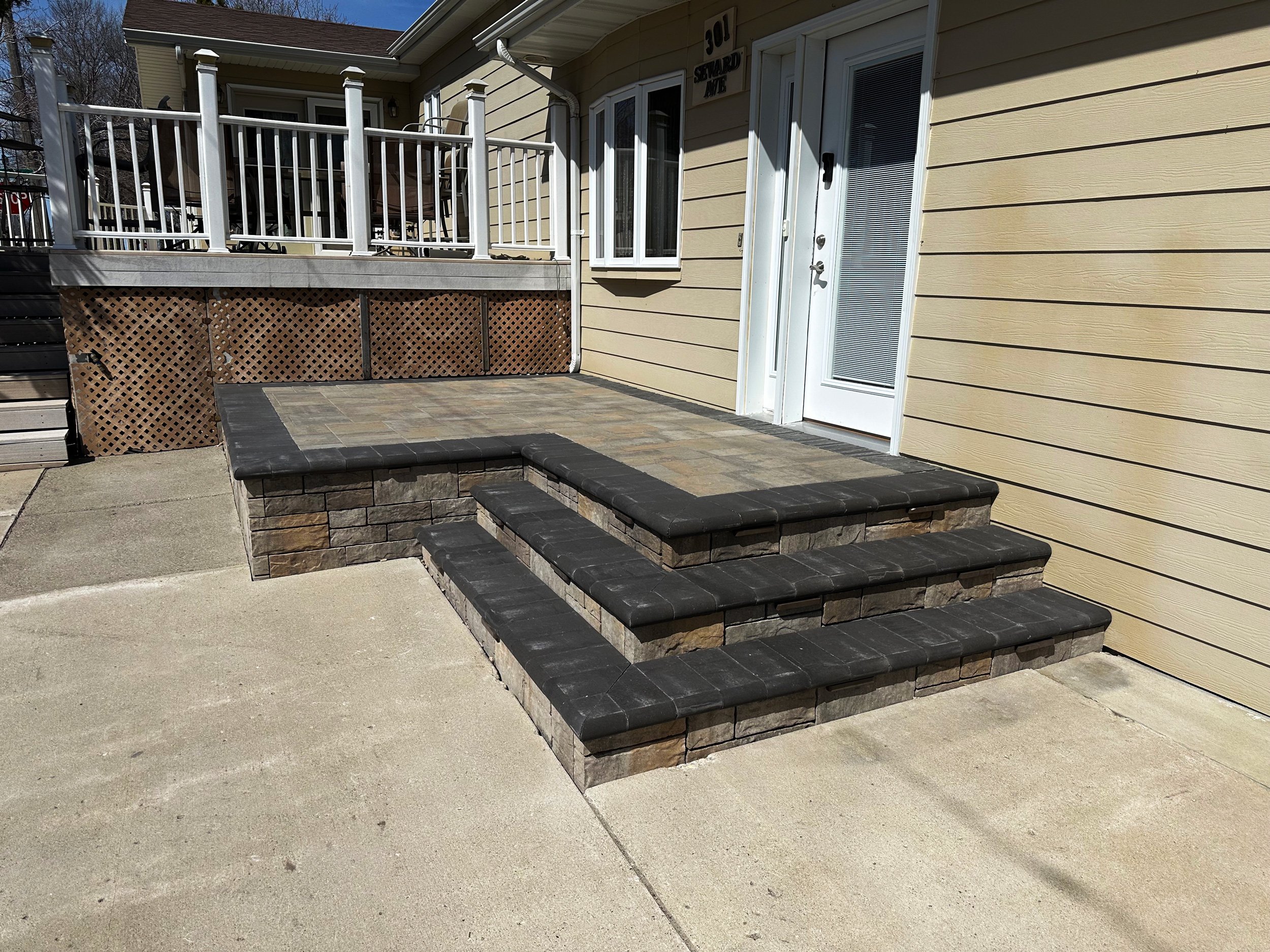 Front porch with black tiled stairs leading up to a beige house with a white door and a small deck with white railing.
