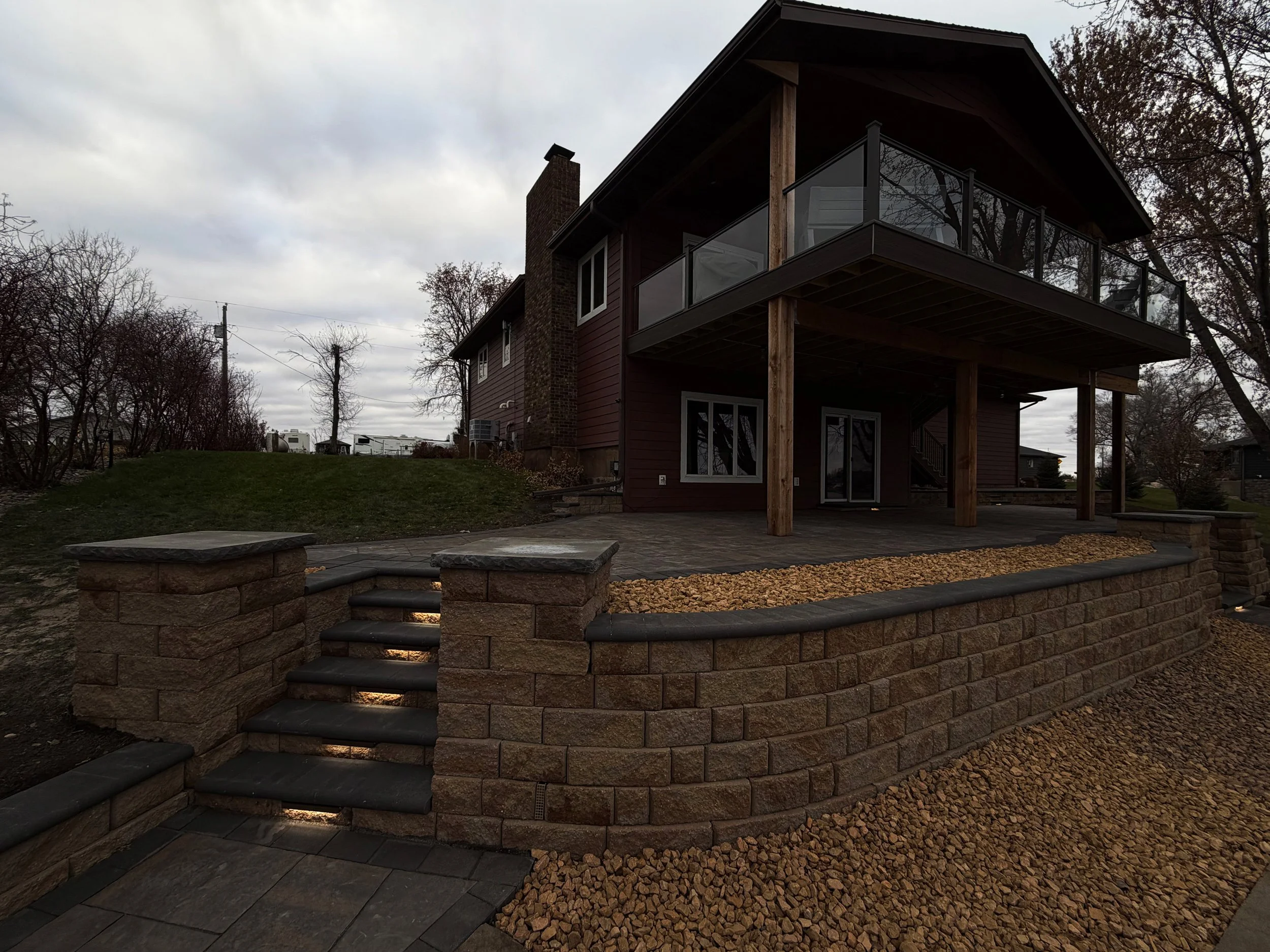 Backyard with a raised patio, stone retaining wall, stairs, and a house with a balcony, surrounded by trees and an overcast sky.