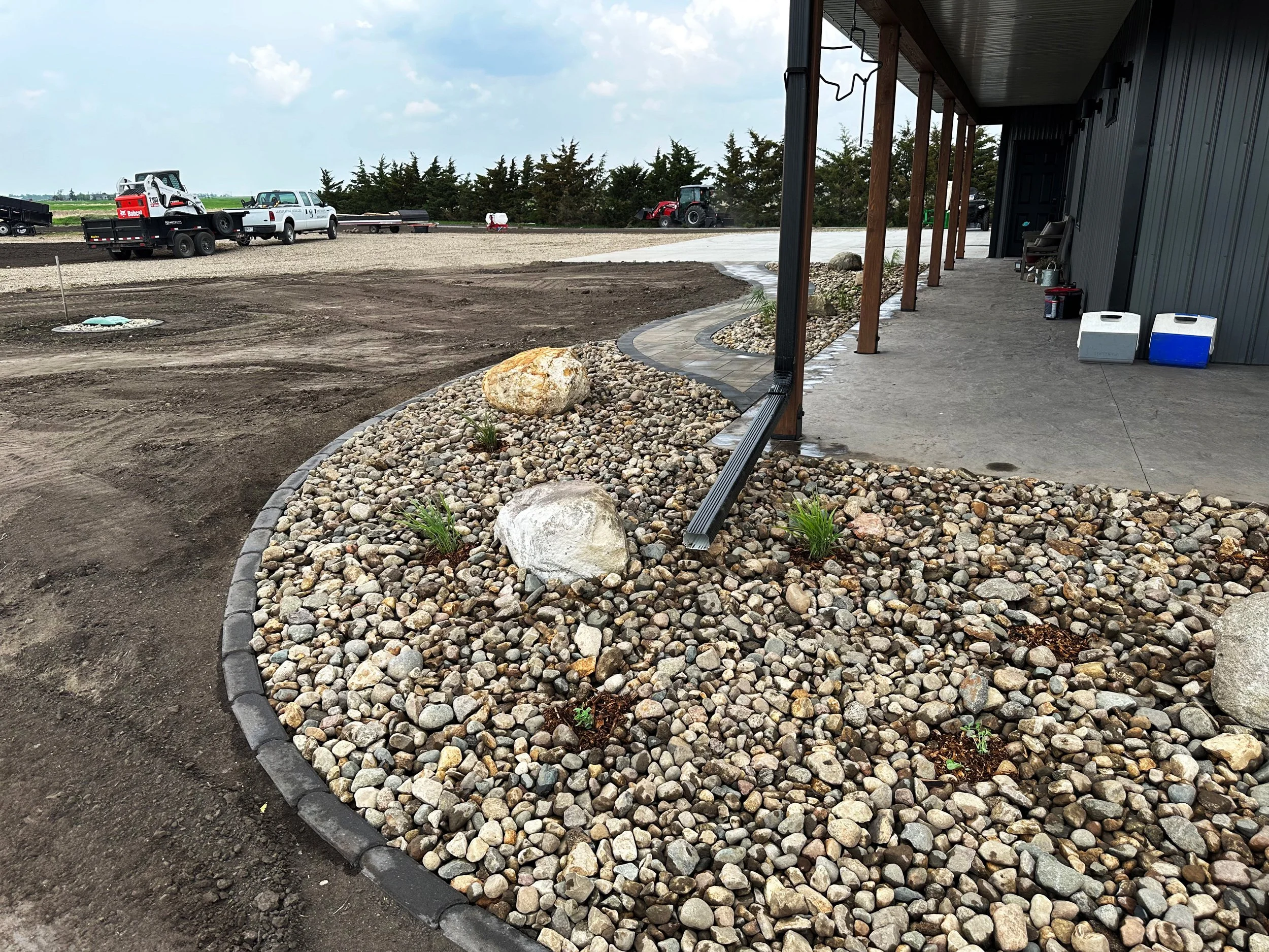 Landscaped area with decorative rocks, small plants, and a curved brick border, near a patio with concrete flooring and a building with a gray exterior.