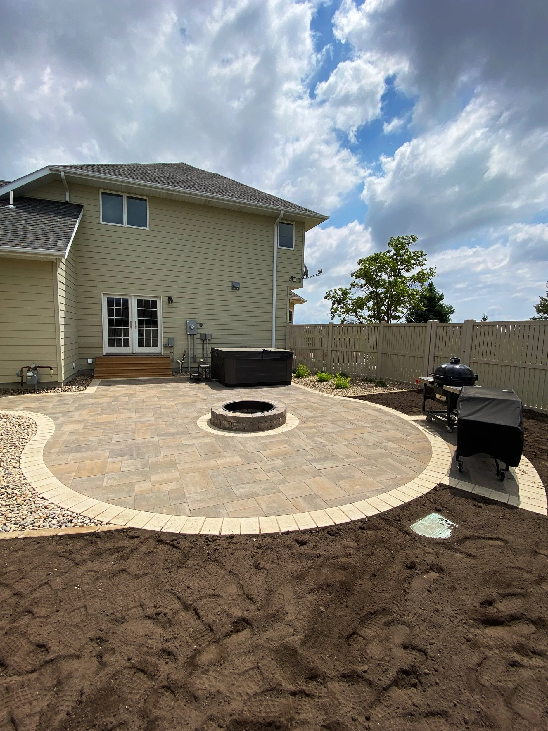 Backyard with newly installed paver patio featuring a central fire pit, surrounded by a wooden fence and outdoor grill, with a two-story beige house and cloudy sky in the background.