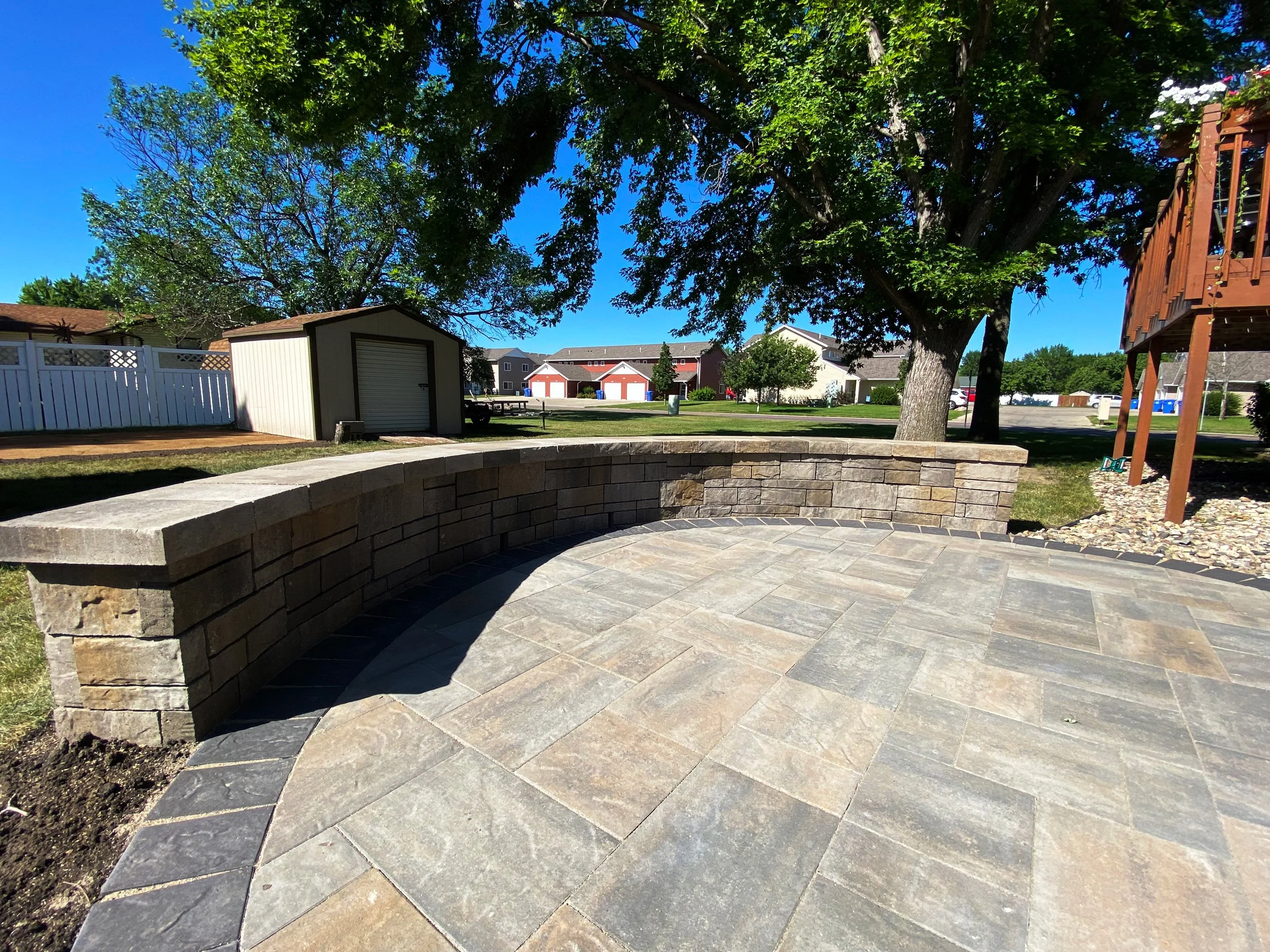 A backyard patio with stone paving and a low curved stone wall. There are large trees, a small shed, and a white fence in the background under a clear blue sky.
