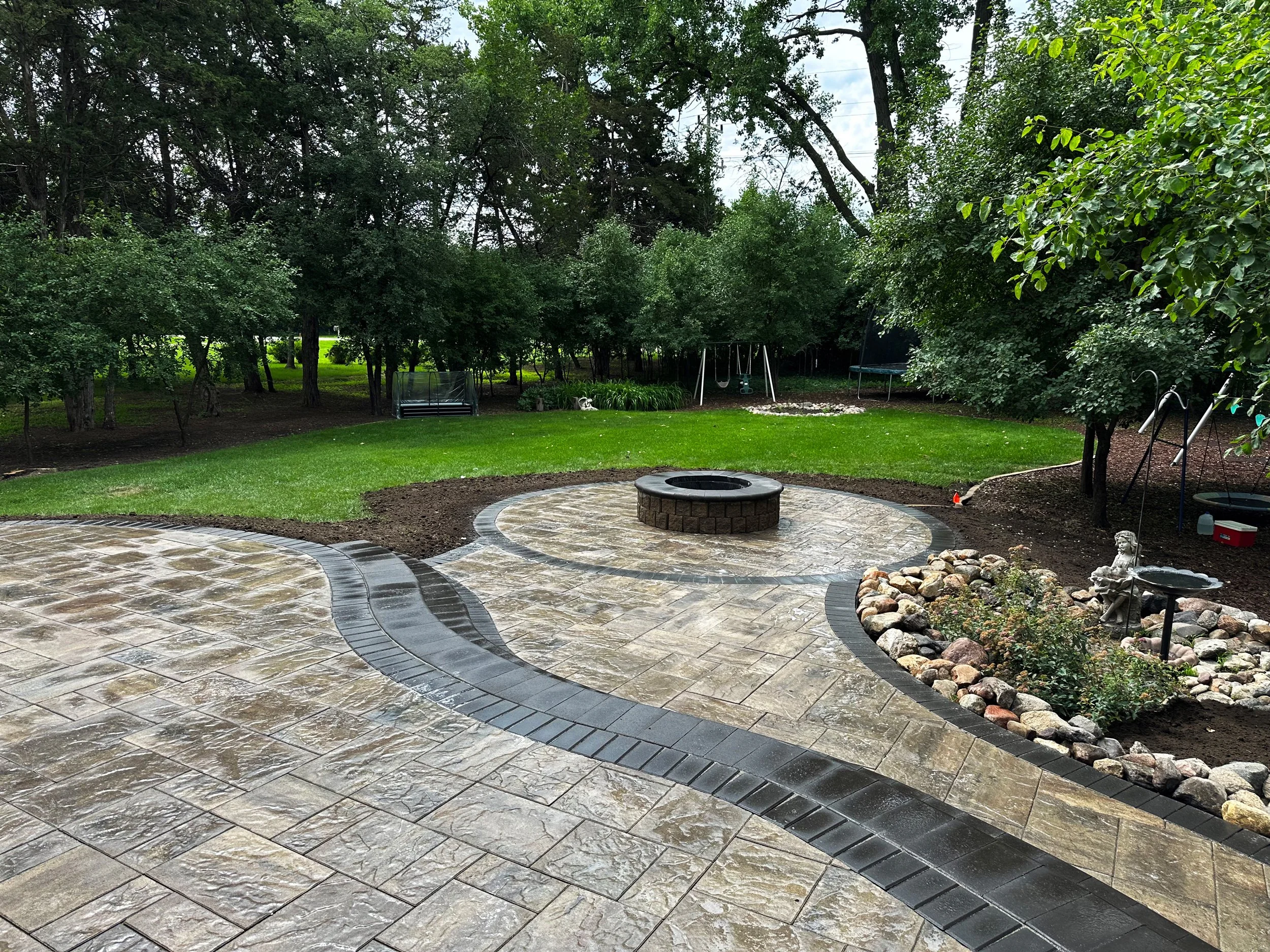 Backyard with stone patio, fire pit, lush green grass, trees, swings, and garden beds.