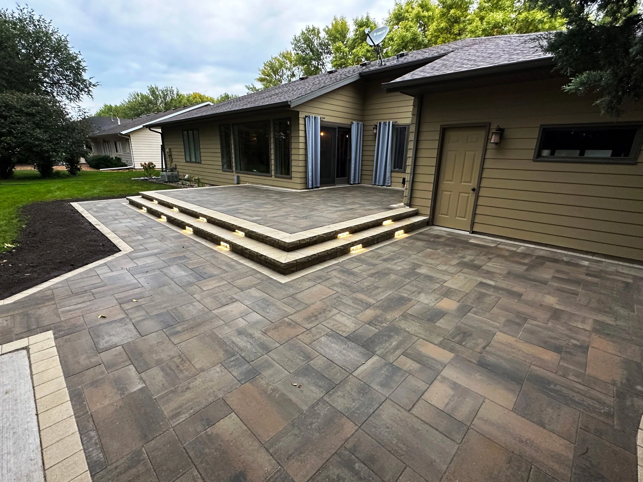 Backyard patio with layered steps, outdoor lighting, and sliding glass doors leading into the house. The area is paved with stone tiles, and there is a grassy lawn with trees surrounding the yard.