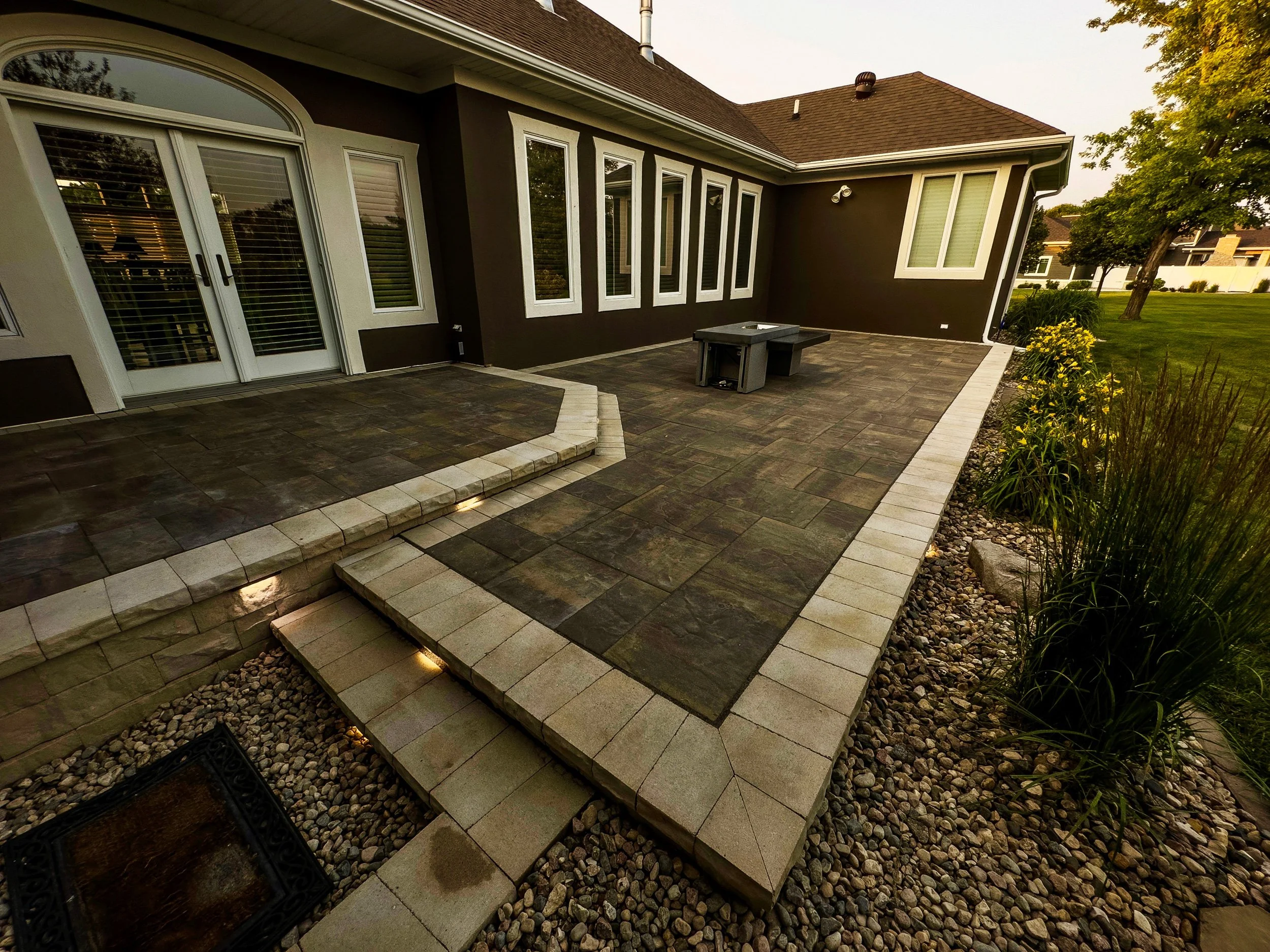 Backyard patio with brick pavers, a step-up area, a small table, and surrounding plants and grass