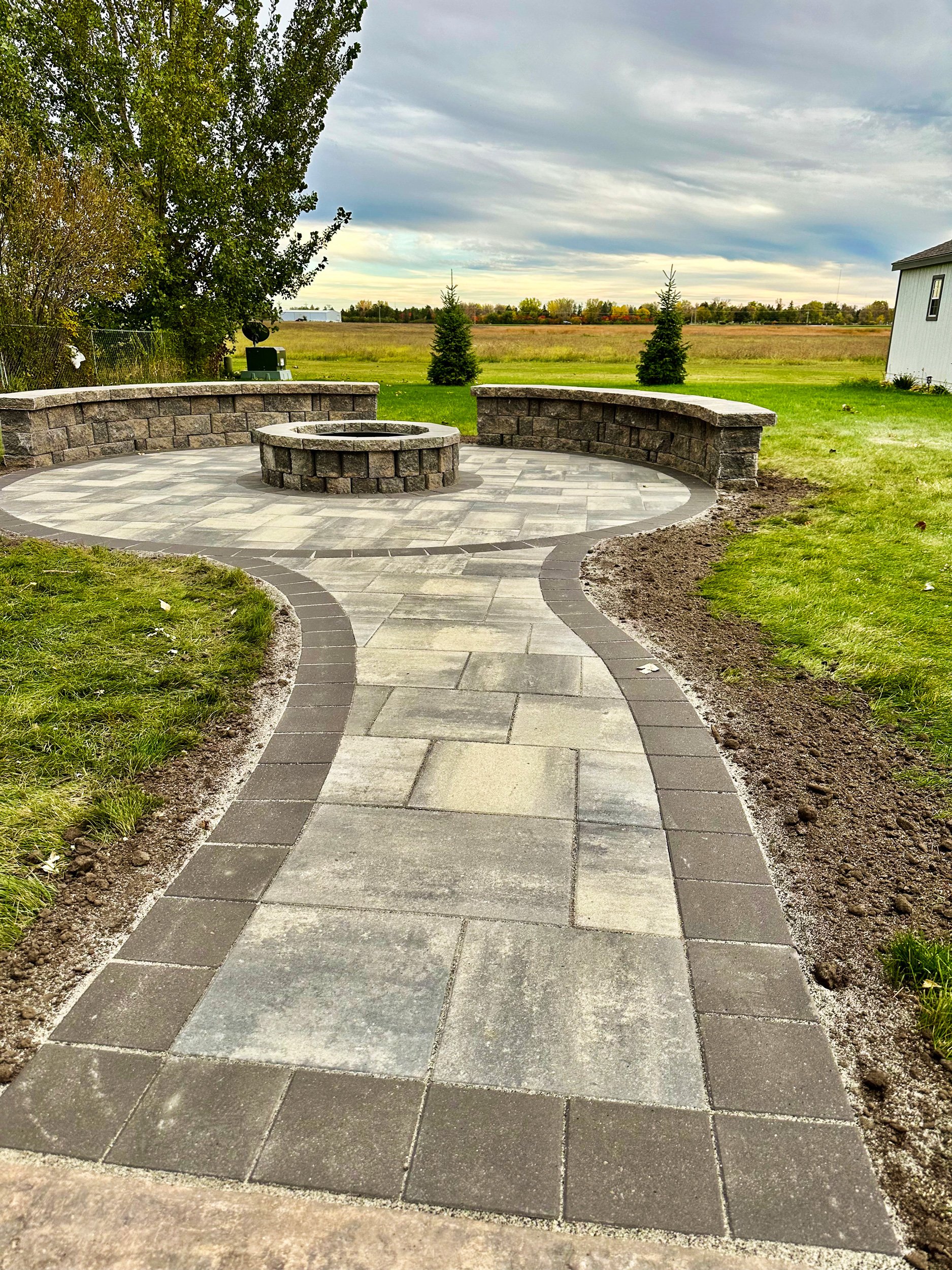 Curved stone and concrete pathway leading to a circular fire pit with a low stone wall in backyard with grassy lawn, trees, and open field under cloudy sky.