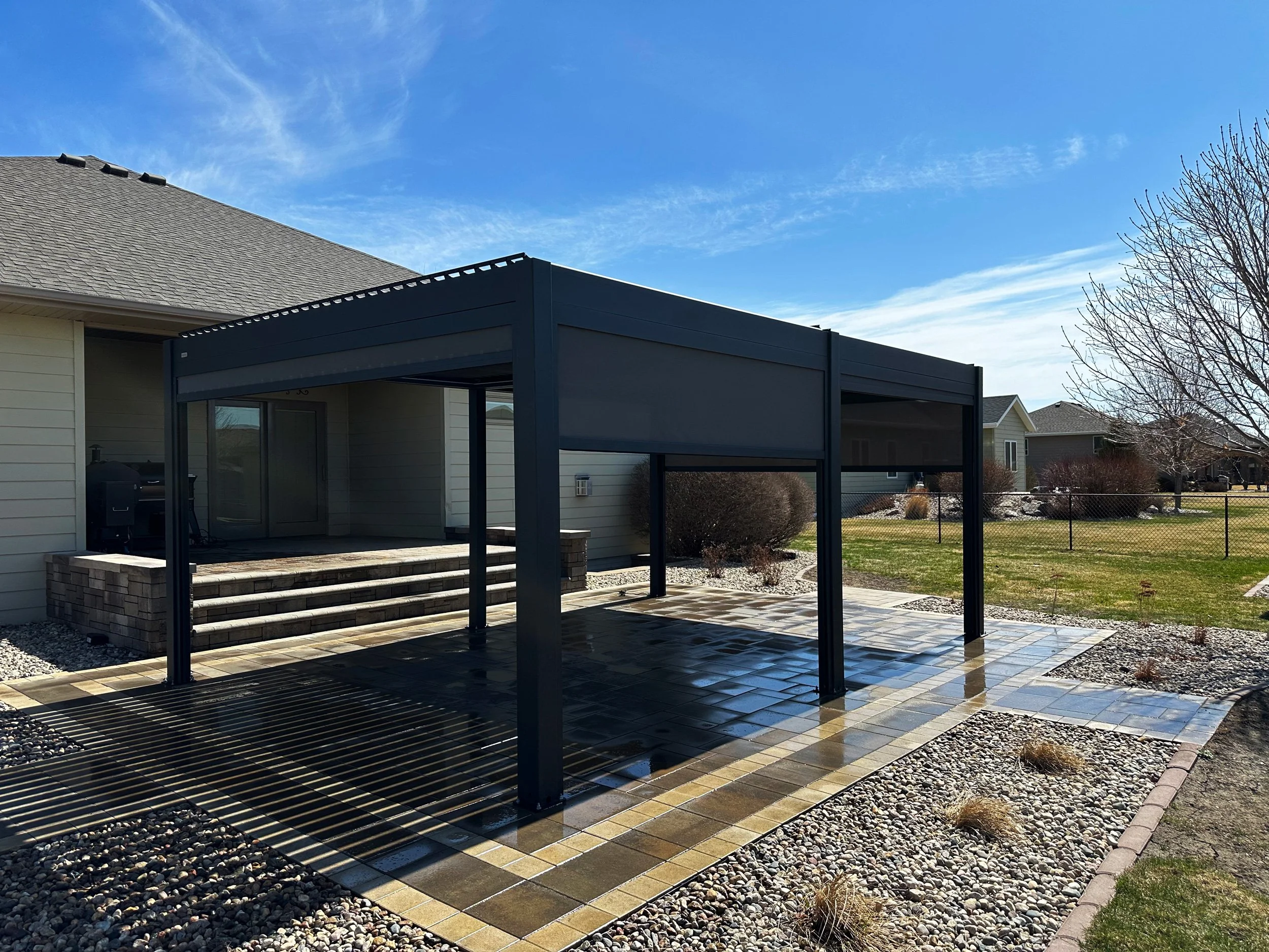 A black metal outdoor pergola on a stone patio in a backyard with grass, shrubs, and trees under a blue sky.