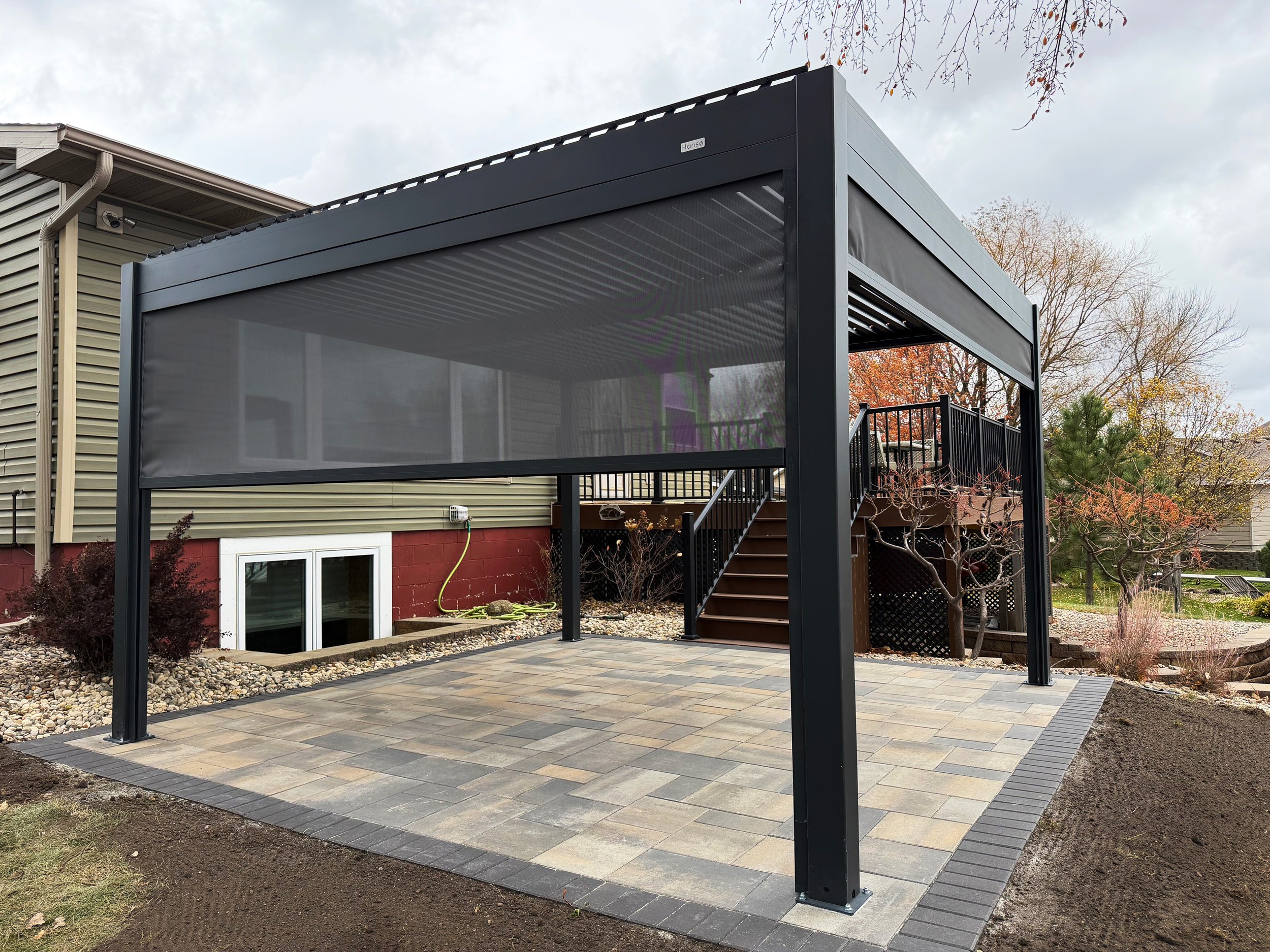 A black metal pergola with a screened roof on a paved patio in a backyard, with stairs leading to an elevated deck and yellow, red, and green trees in the background.