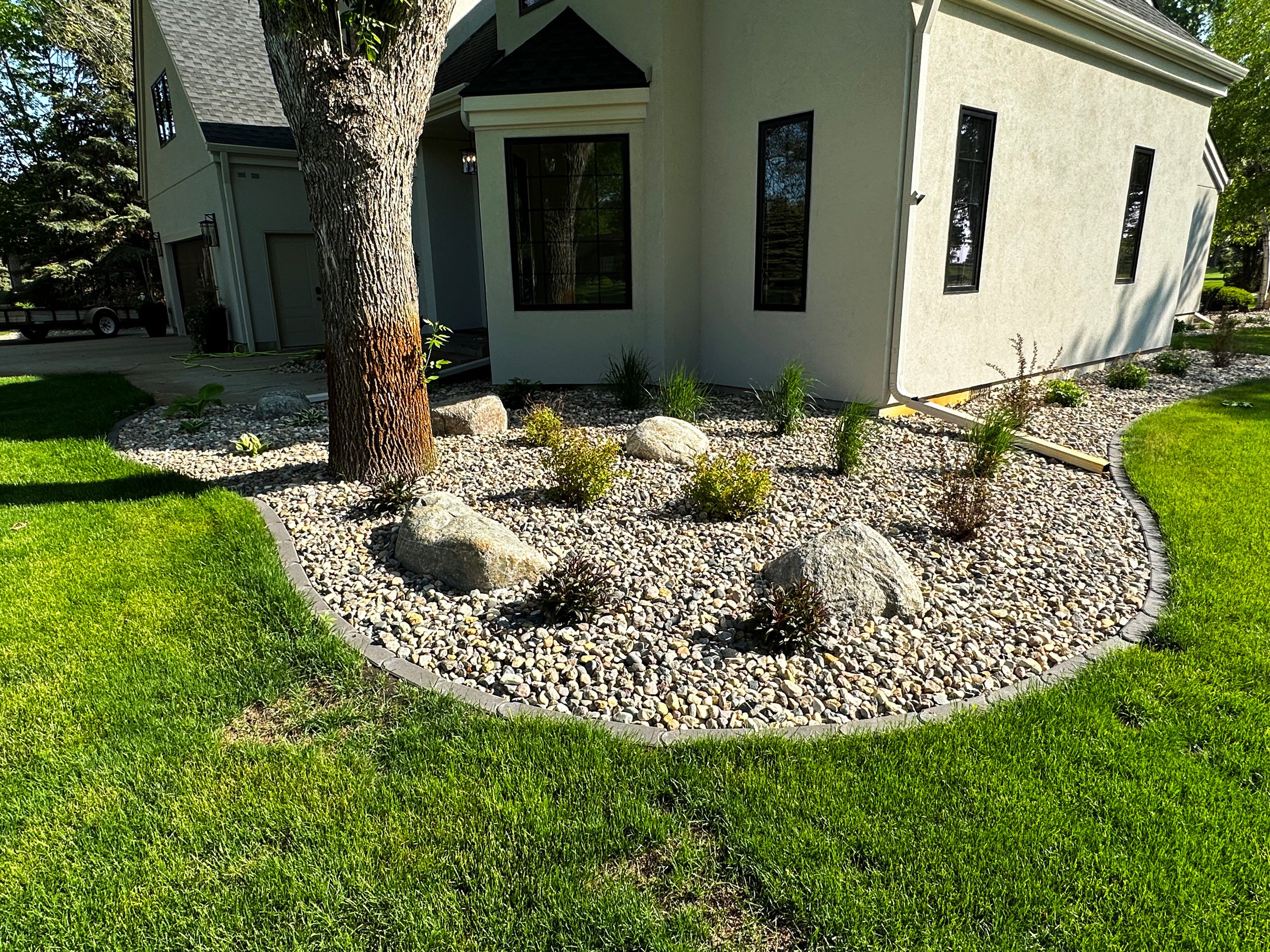 Landscaped front yard with a tree, rocks, shrubs, green grass, and a house with beige exterior and black windows.
