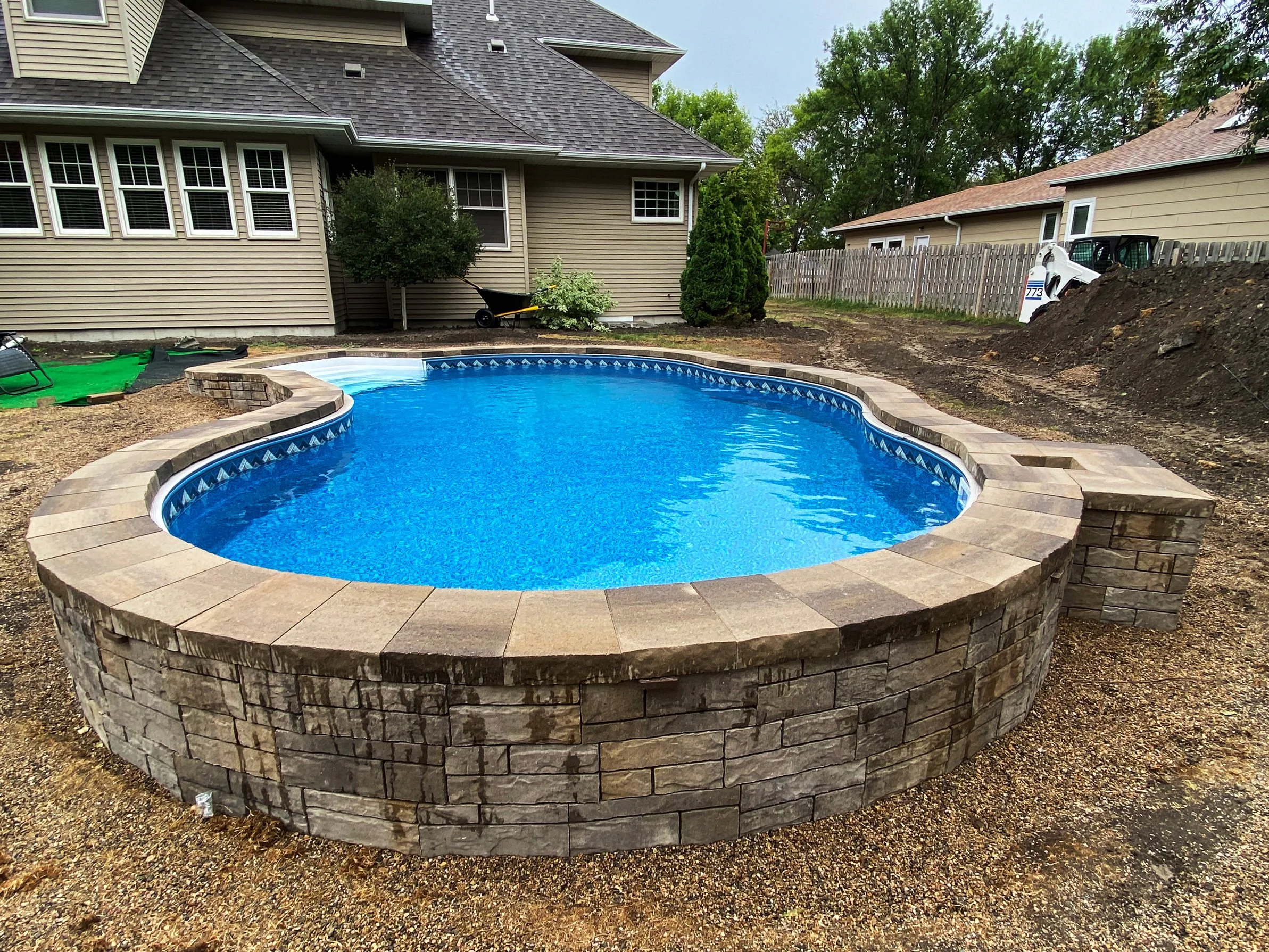 In-ground backyard swimming pool with stone coping and a blue interior, under construction with dirt and gravel surrounding it, in a suburban backyard with house and trees in the background.