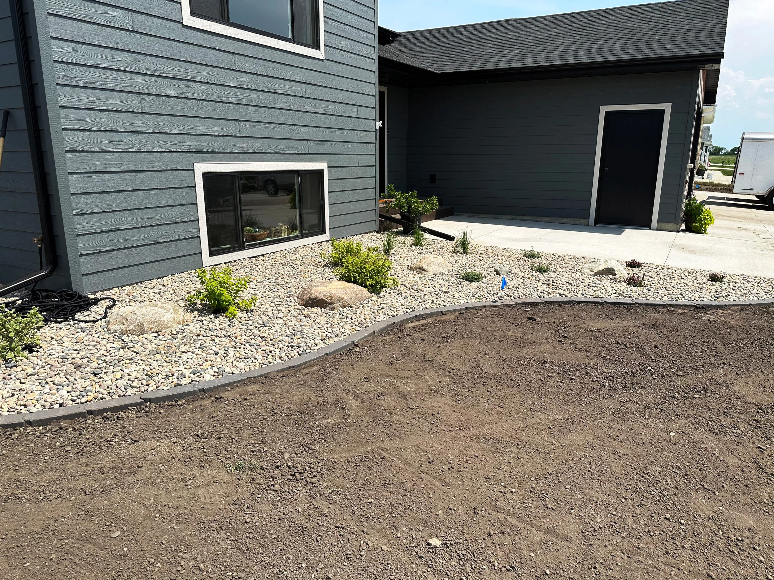 A landscaped front yard with a gravel bed, small green bushes, and dirt area in front of a dark blue house with white trim. There is a concrete sidewalk and a black door on the house.