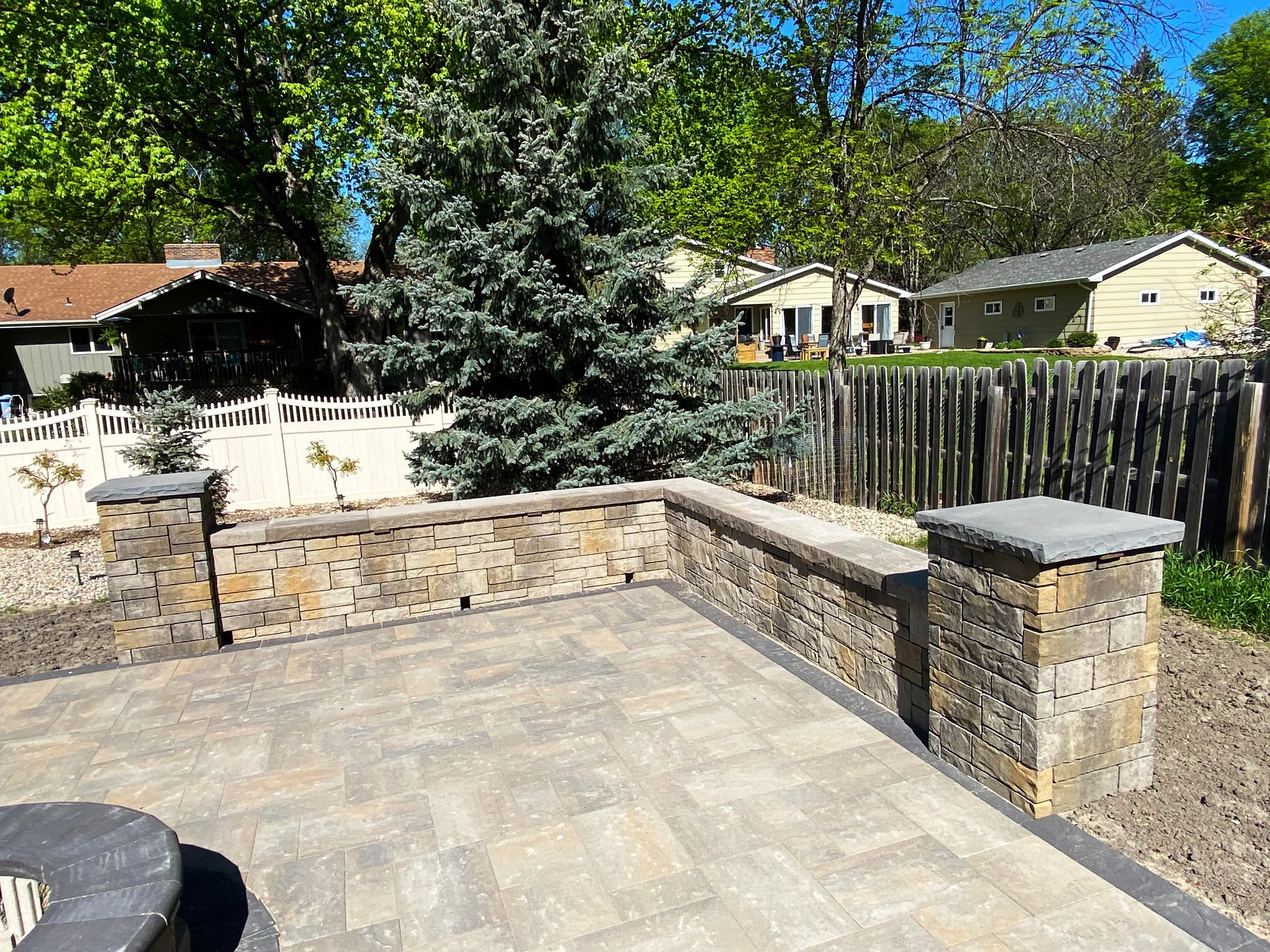 A backyard patio with a low stone wall, surrounded by trees and neighboring houses under a blue sky.