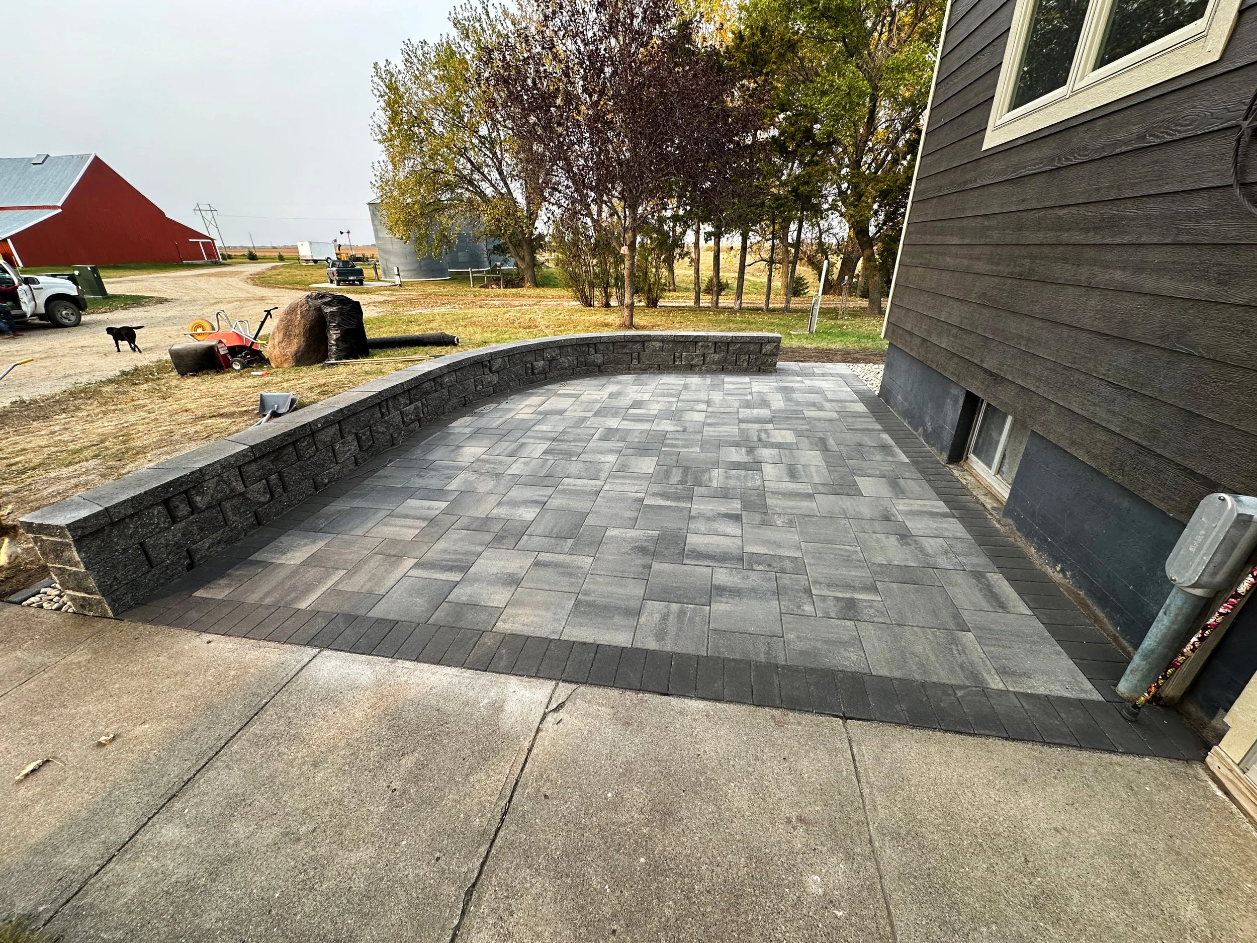 A newly paved outdoor patio area with gray stone tiles, bordered by a black stone edging, next to a dark brown house with white-trimmed windows. The patio is set against a backdrop of trees and farm buildings, with some lawn and yard equipment visibl