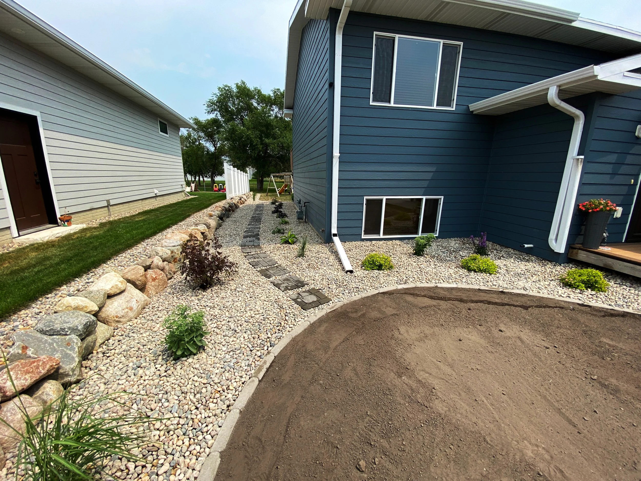 Backyard with gravel landscaping, small plants, a curved stone pathway, a blue house, and a neighboring house with a brown door, and a grassy area in the background.