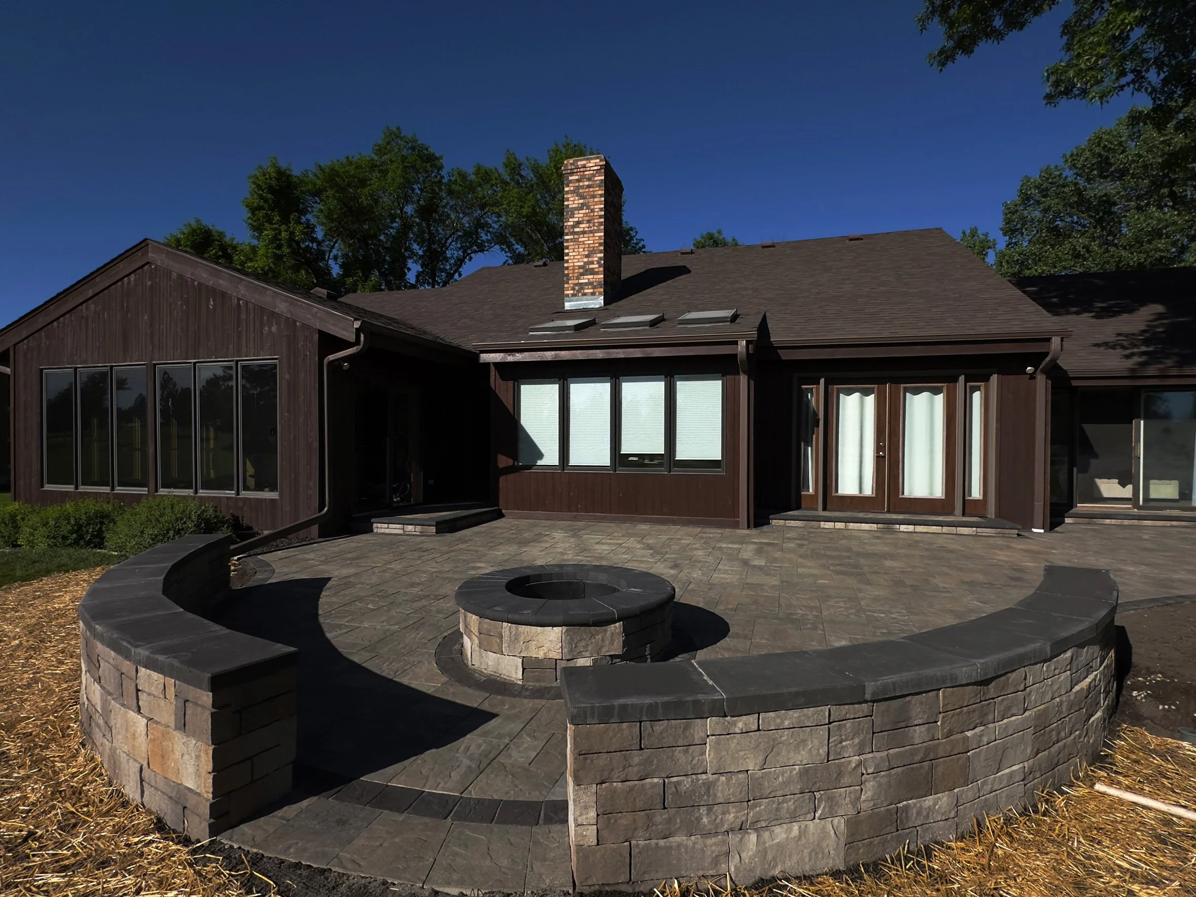 Backyard patio with a circular fire pit, stone seating wall, and a brown house with large windows and a brick chimney.