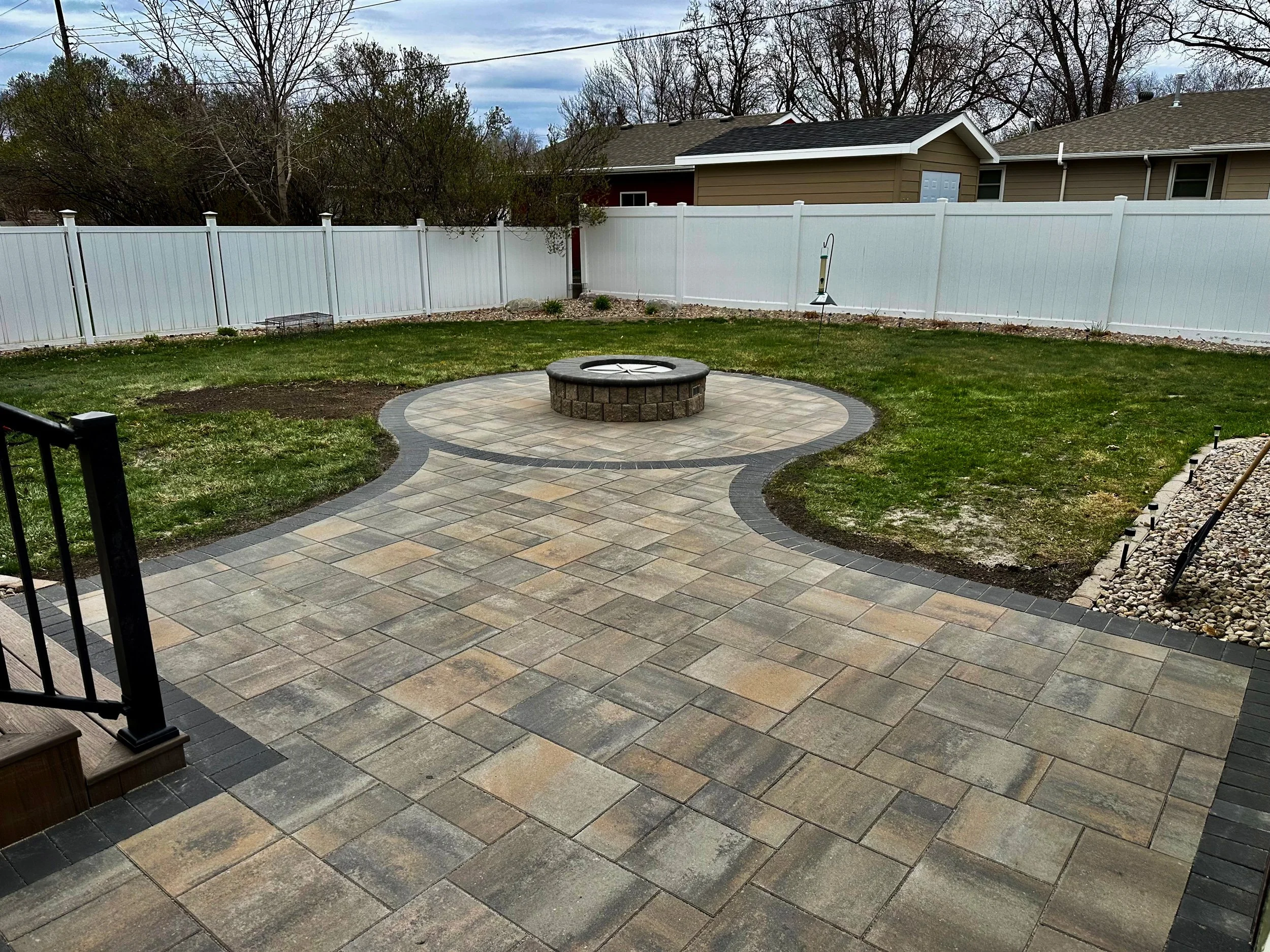 Backyard with a stone patio, fire pit in the center, grass lawn, white fence surrounding the yard, and neighboring houses in the background.