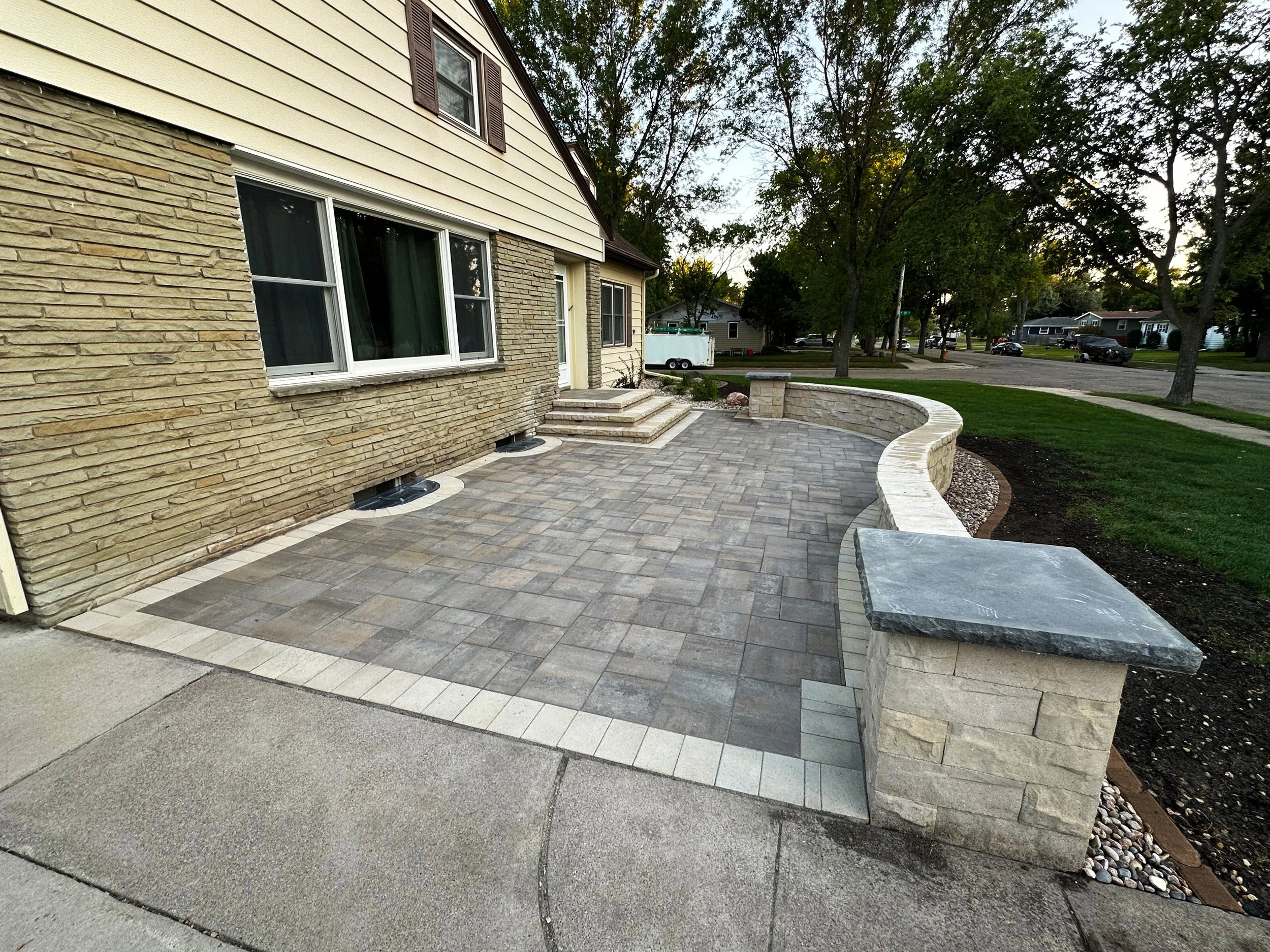 Backyard patio with stone pavers, low stone wall, stairs, and a bench, adjacent to a house with brick and siding exterior, a large window, steps leading to the door, and surrounding trees and grass.