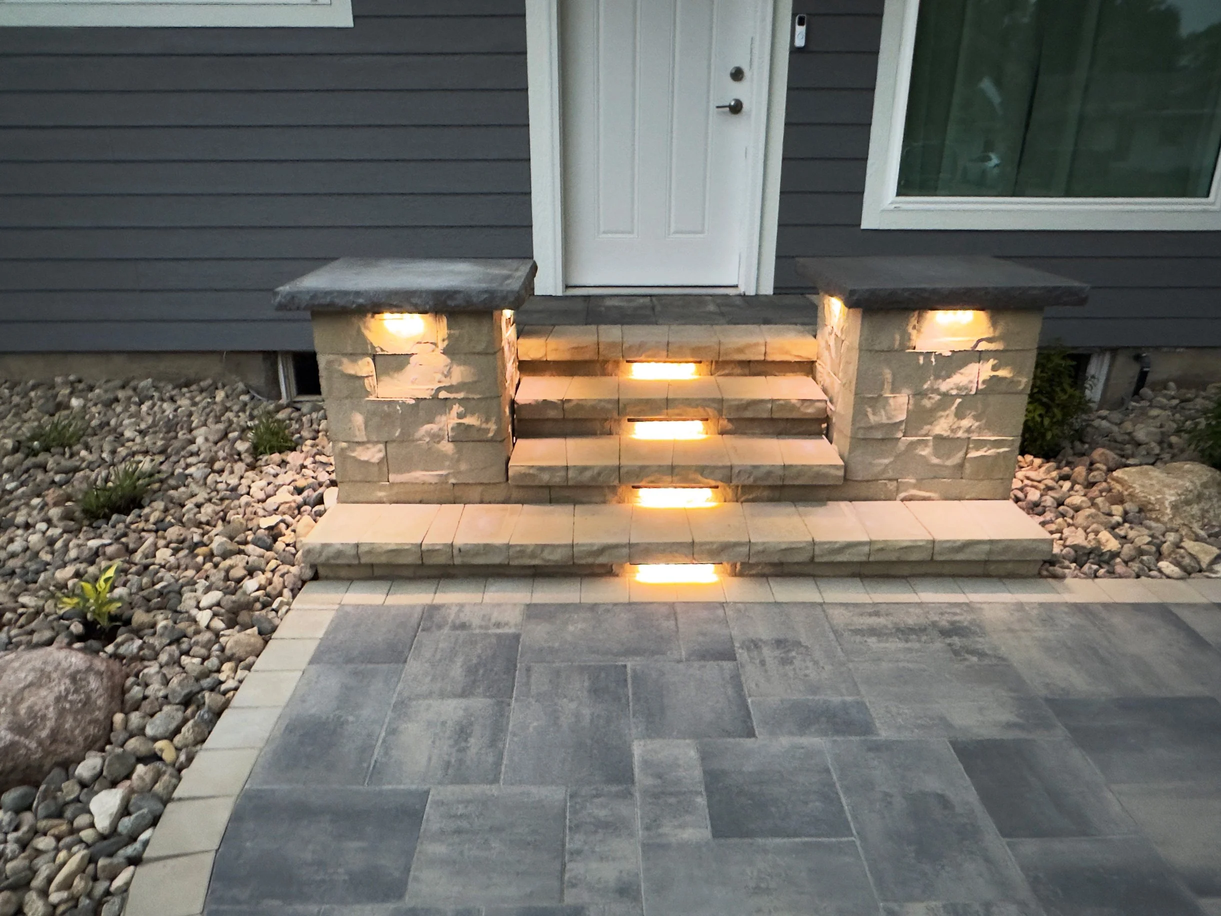 Exterior front porch with stone steps lit by built-in lights, gray house siding, white door, and window.