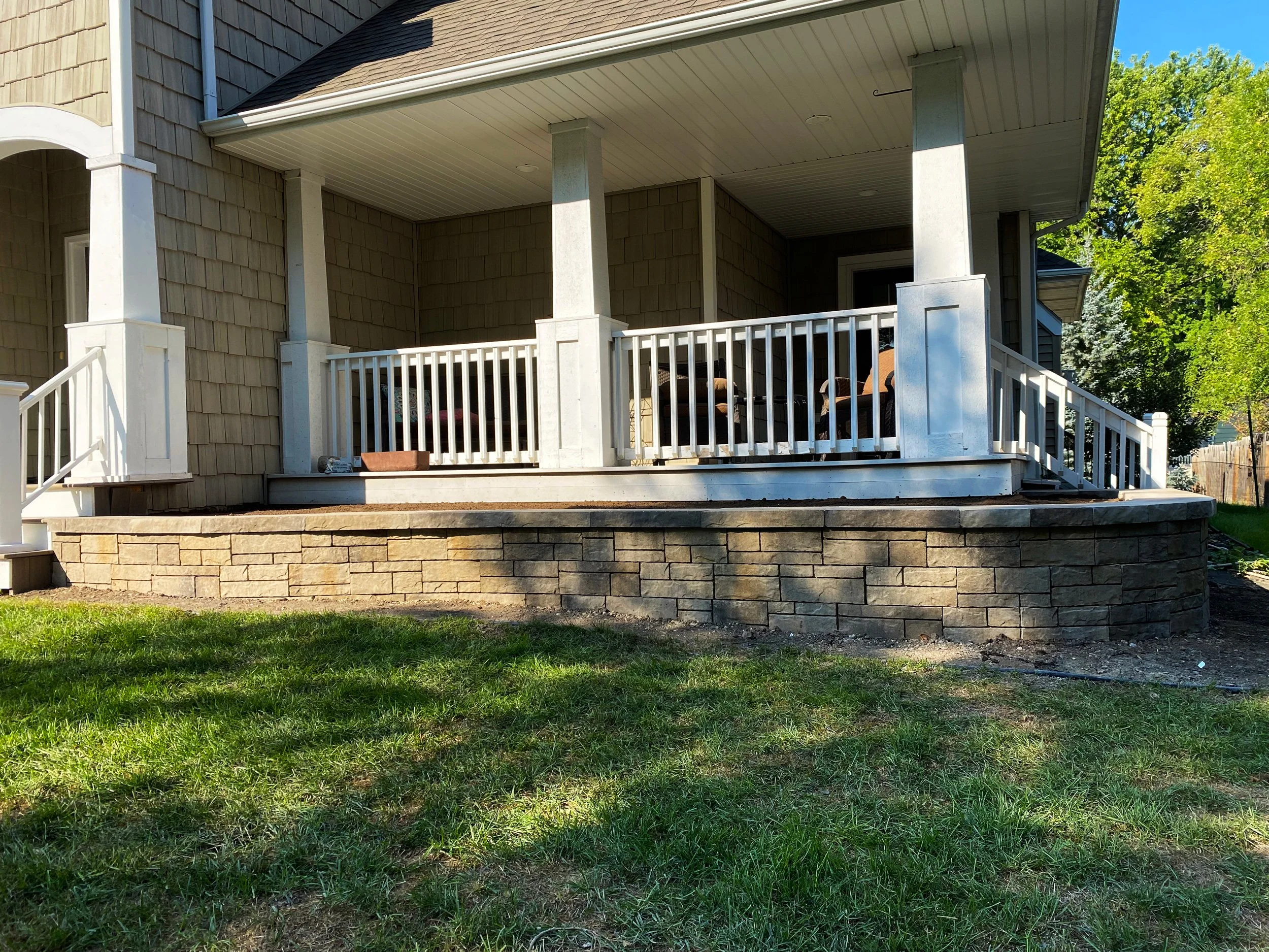 A house with a wooden exterior, a raised porch with white railing, stone foundation, and some outdoor furniture, under a blue sky with green trees in the background.
