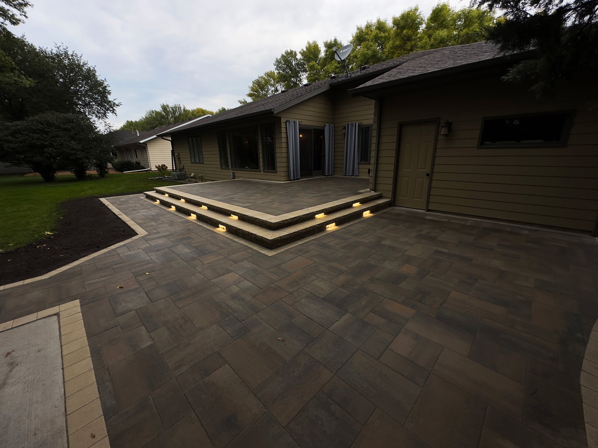 Backyard patio with steps and a house in the background, landscaped with trees and grass, during daytime.