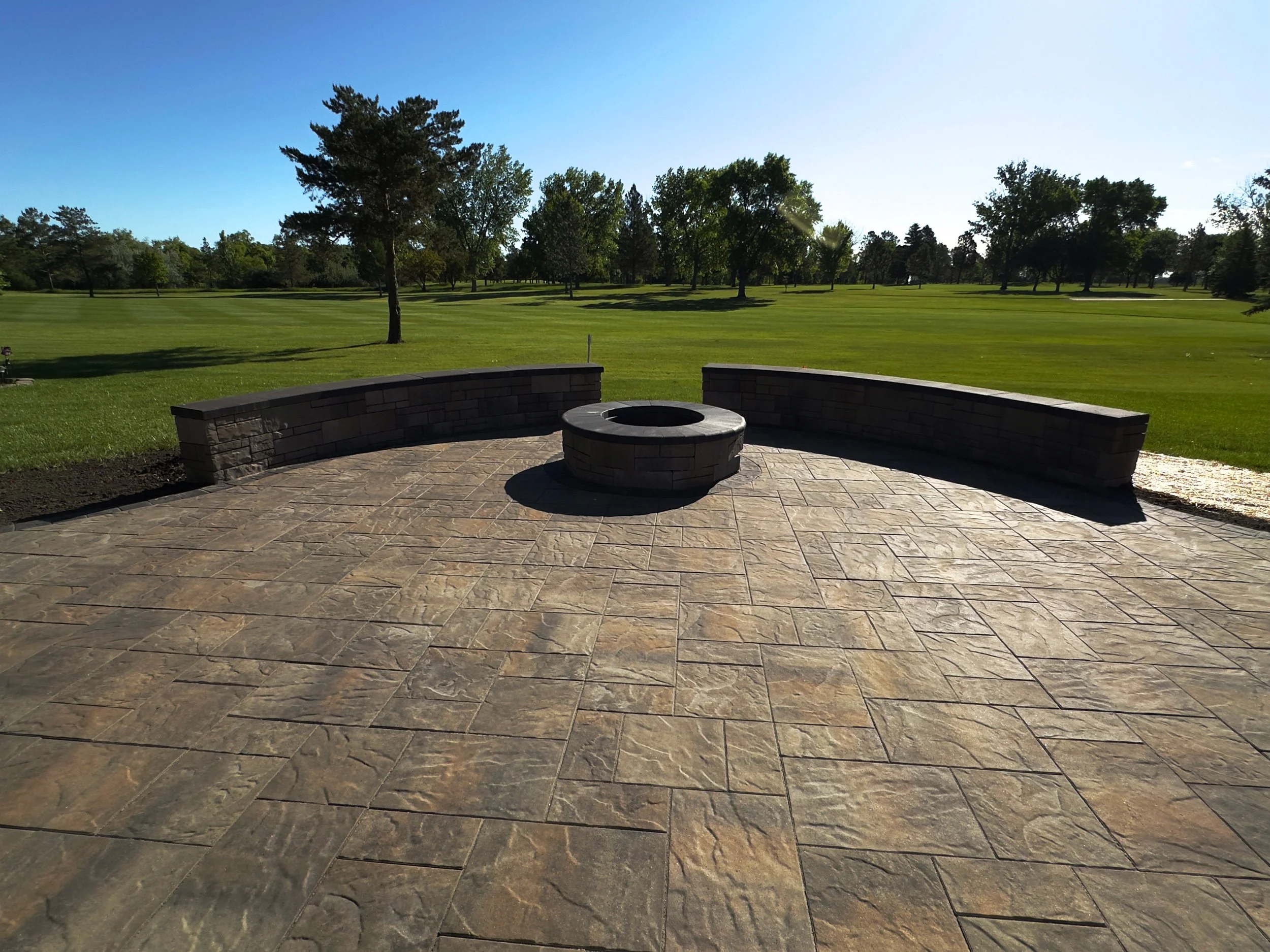 A stone patio with a fire pit surrounded by a curved stone wall, overlooking a green golf course with trees and a clear blue sky.