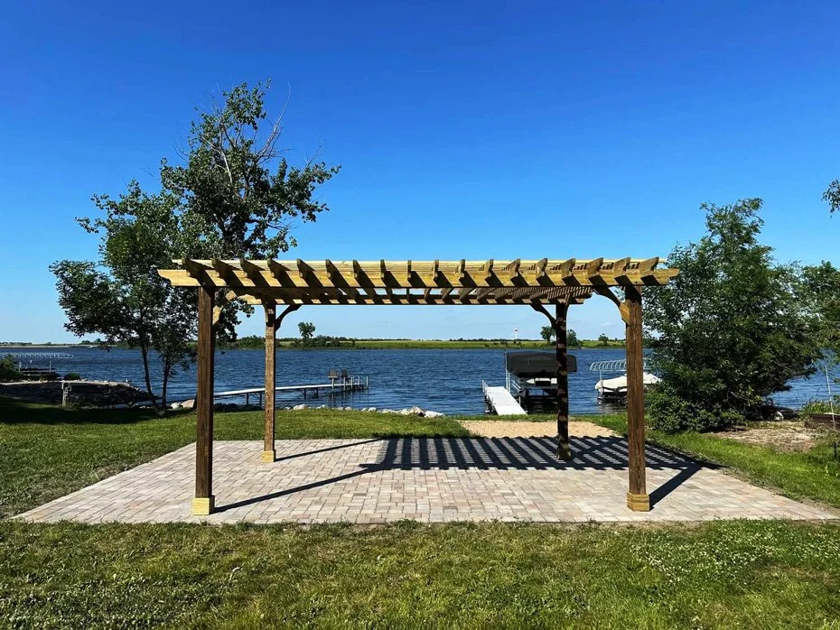 A wooden pergola with a slatted roof over a brick patio near a body of water with trees and boats in the background.