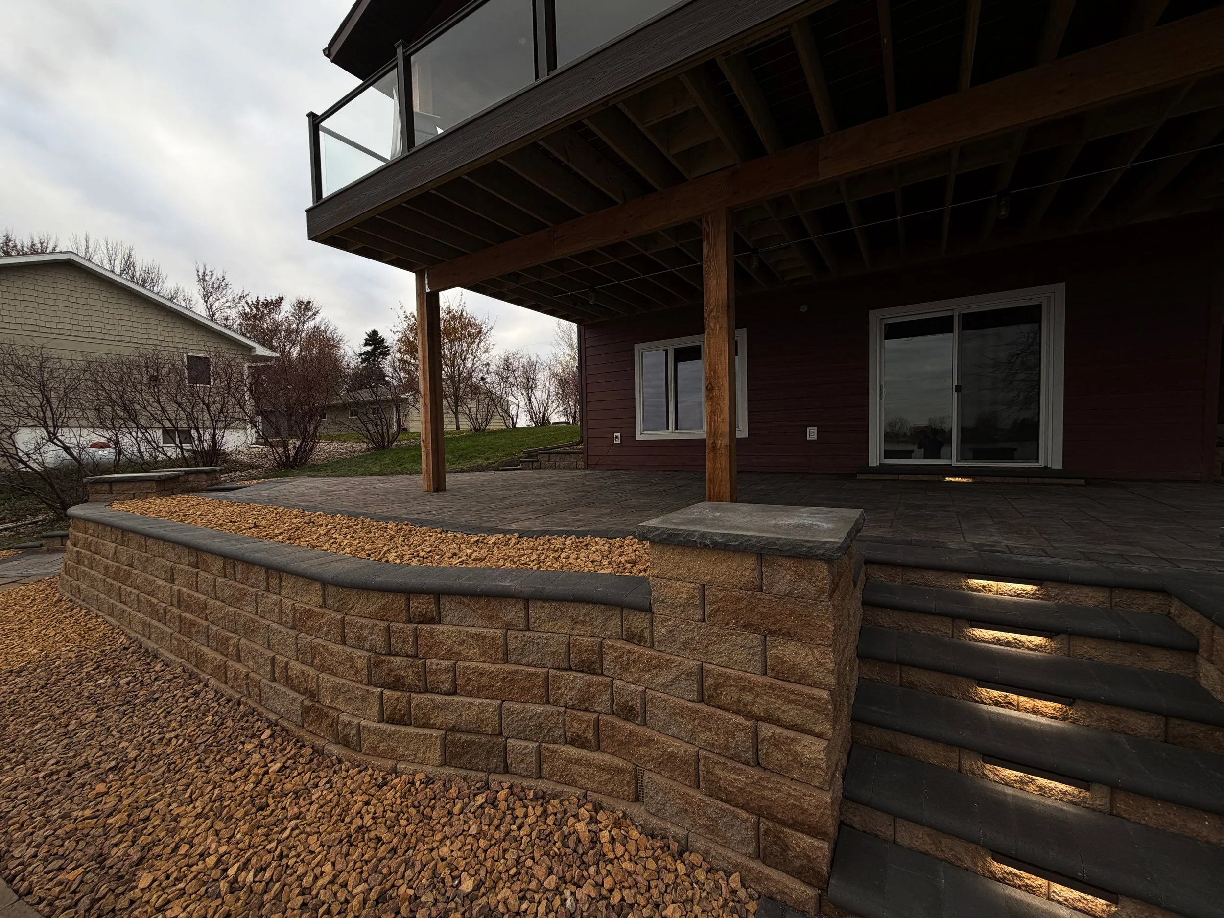Backyard patio with stone wall and steps leading up to a house with a wooden deck and sliding glass door, under an overcast sky.