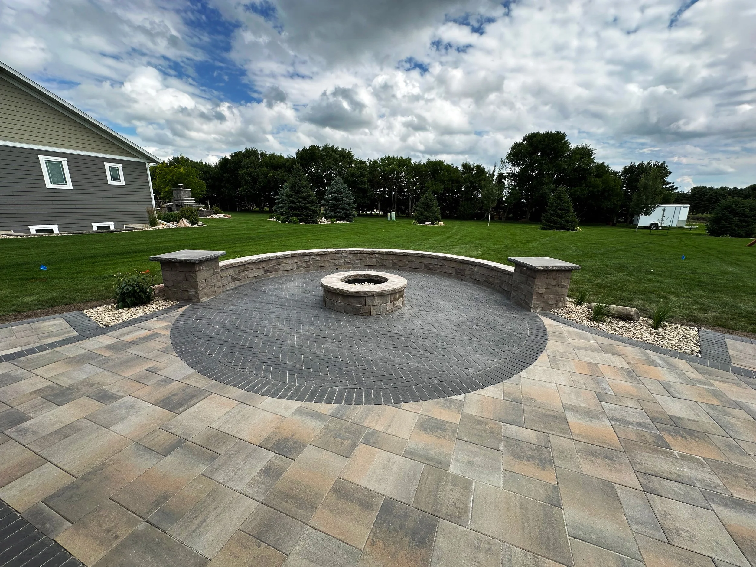 Backyard with a circular brick patio, a fire pit in the center, and a stone bench wall surrounding part of the patio. Green grass and trees in the background, with partly cloudy sky.
