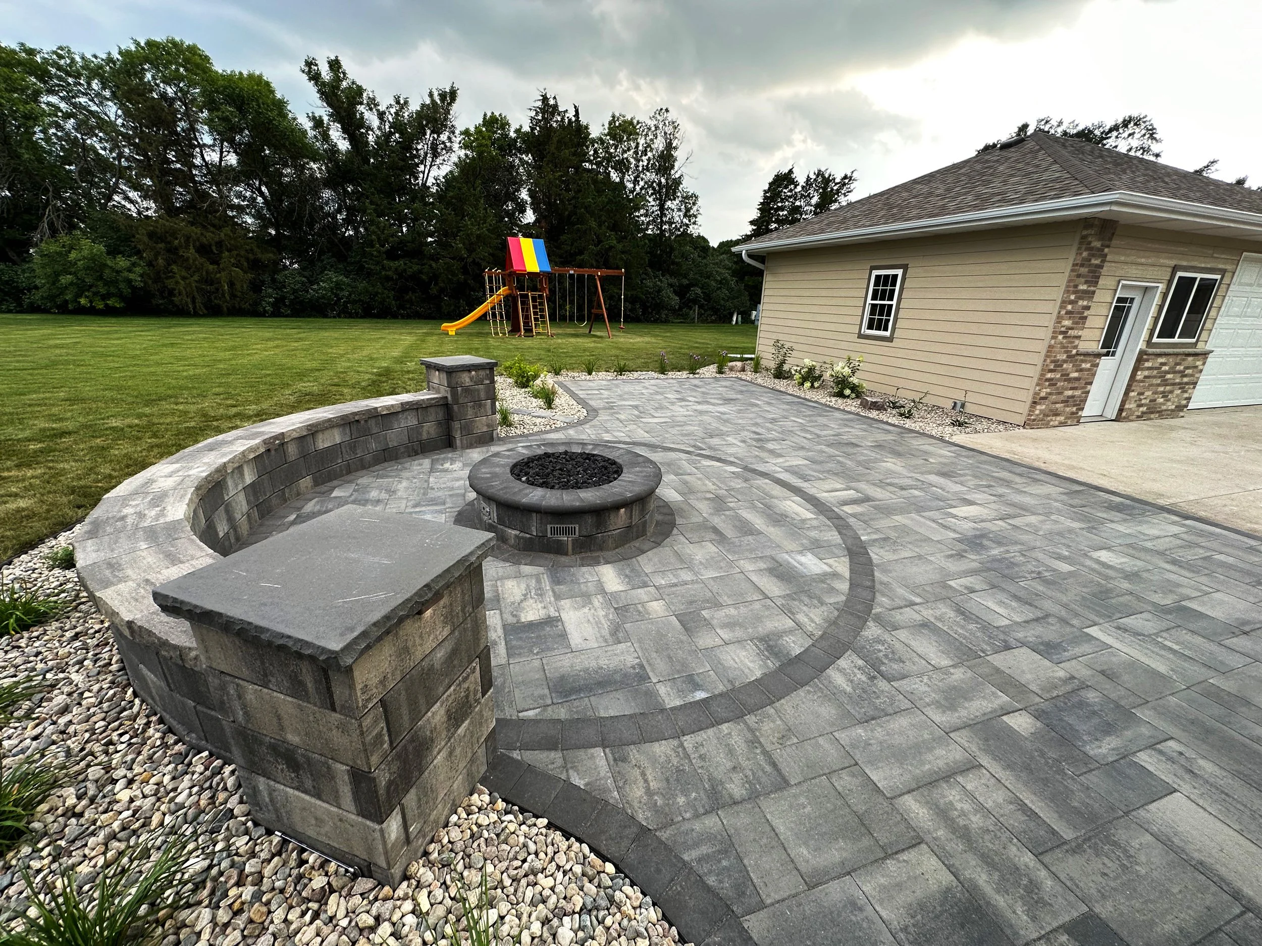 This image shows a backyard patio area with gray brick pavers, a fire pit, and a curved stone bench. There are small plants and white rocks along the edge. In the background, there is a grassy lawn, a children's playground with a slide, swing set, an