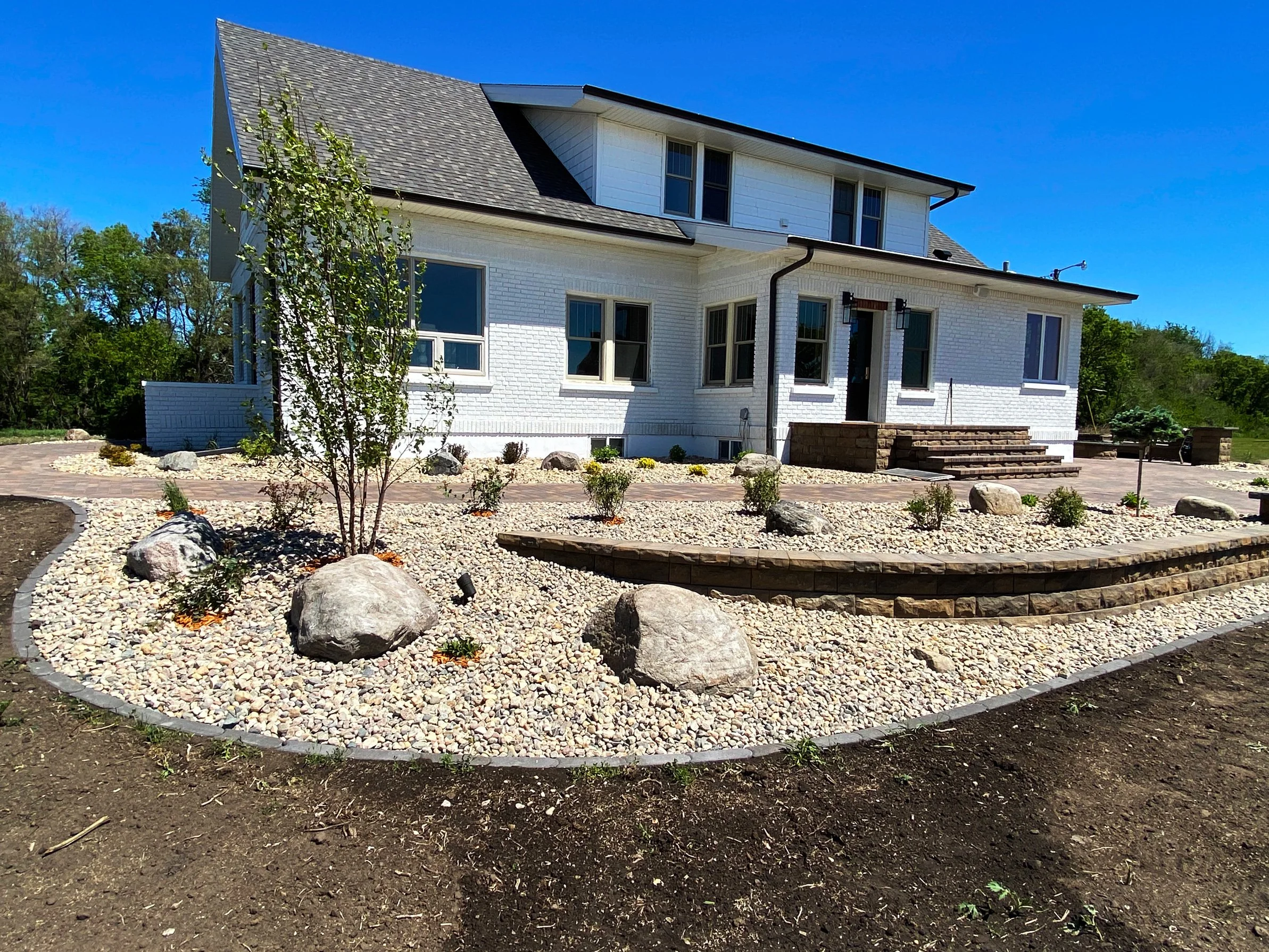 A two-story white house with a dark roof and large windows, surrounded by a landscaped yard with rocks, small plants, and a paved pathway under a clear blue sky.