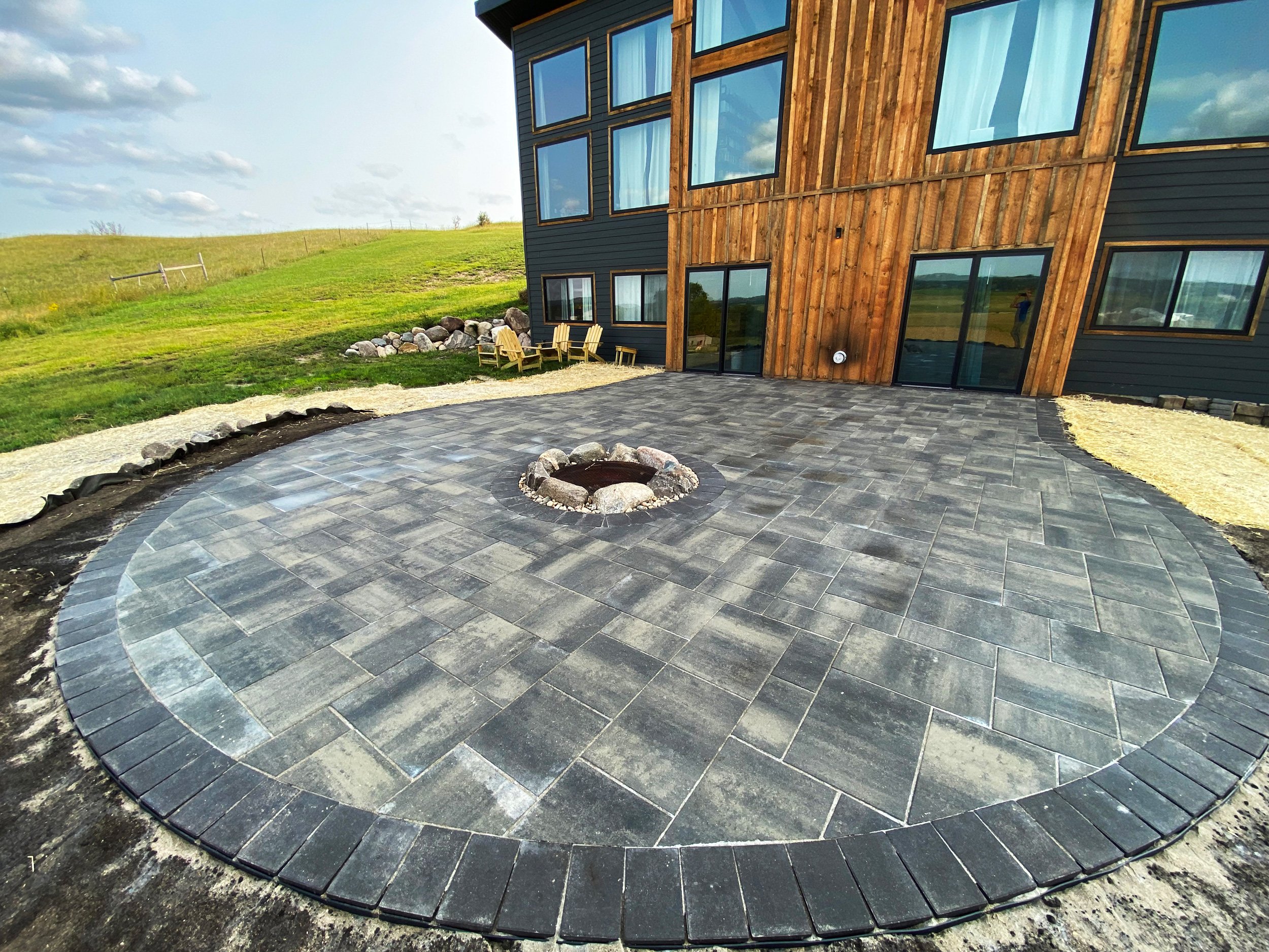 Newly paved circular patio with a fire pit in the center, adjacent to a modern house with large windows and wood siding, surrounded by a grassy yard and open landscape.