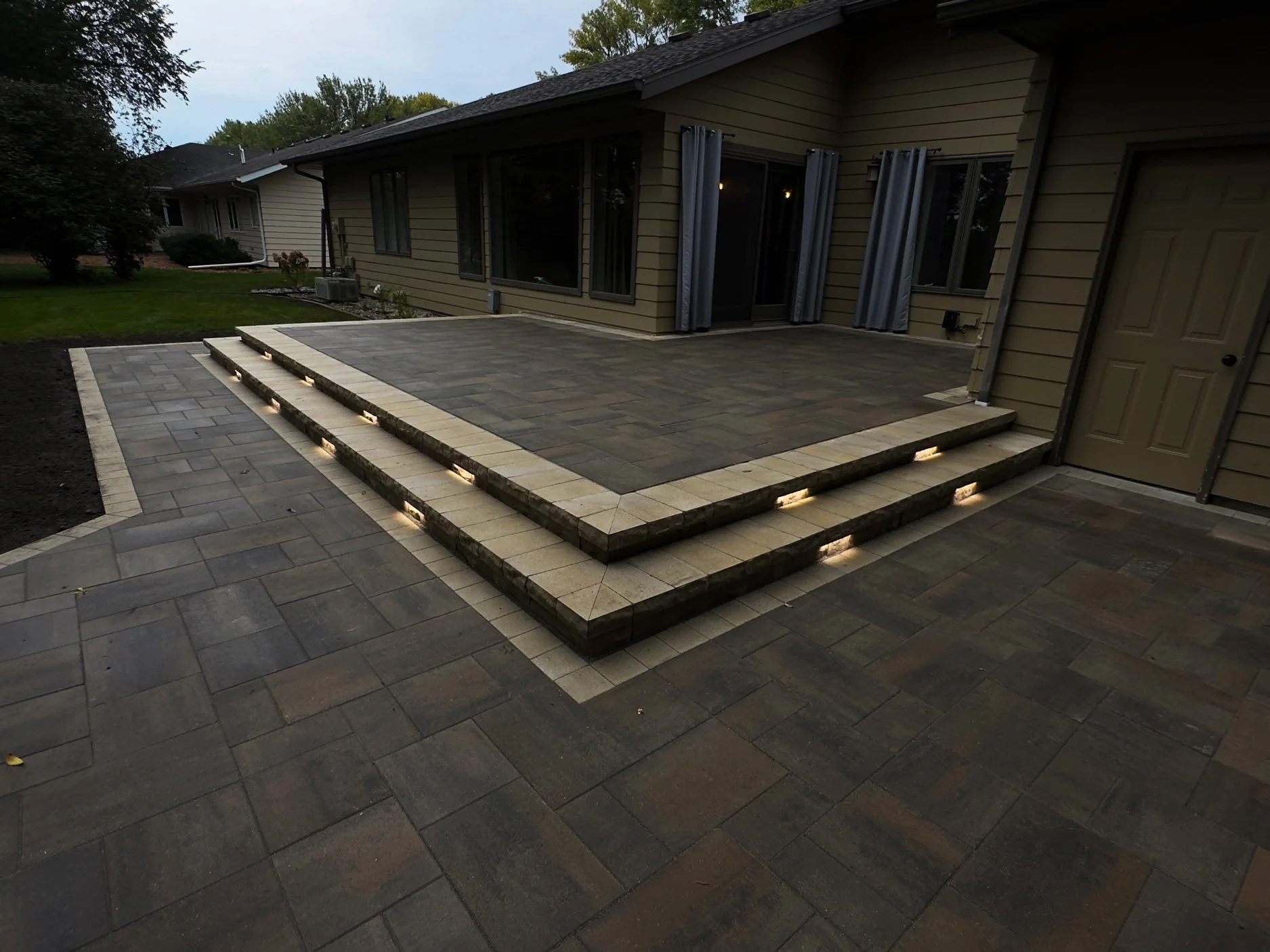 A backyard patio with a multi-level stone design, featuring steps with built-in lighting, adjacent to a house with doors and windows, and a lawn with trees in the background.